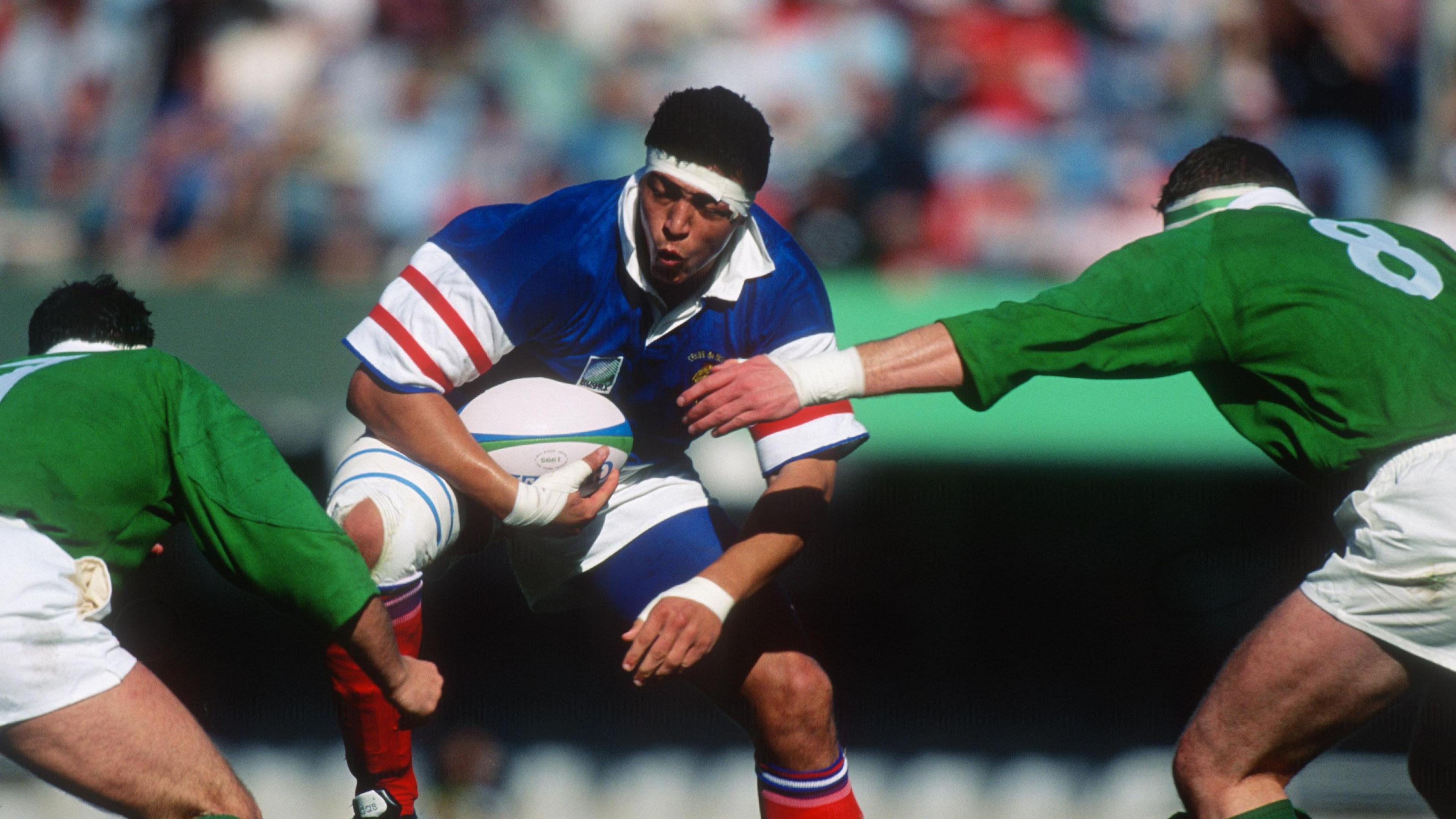 Abdelatif Benazzi wearing blue French shirt and white headband carries the ball and jumps into contact with two Irish players in green at the 1995 Rugby World Cup in South Africa