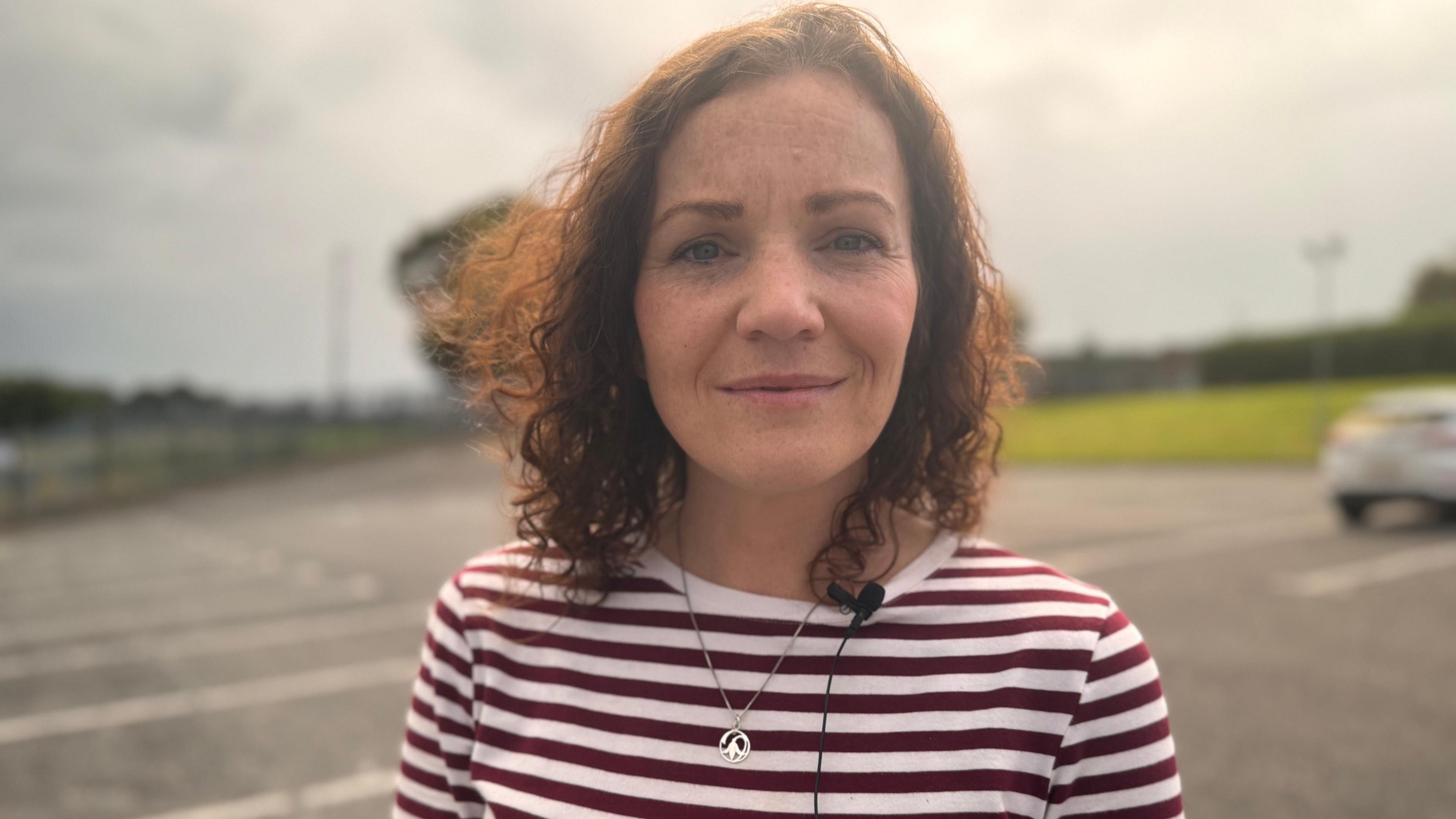 A woman with brown curly hair is looking into the camera. She's wearing a silver necklace and stripped top and is standing outside.