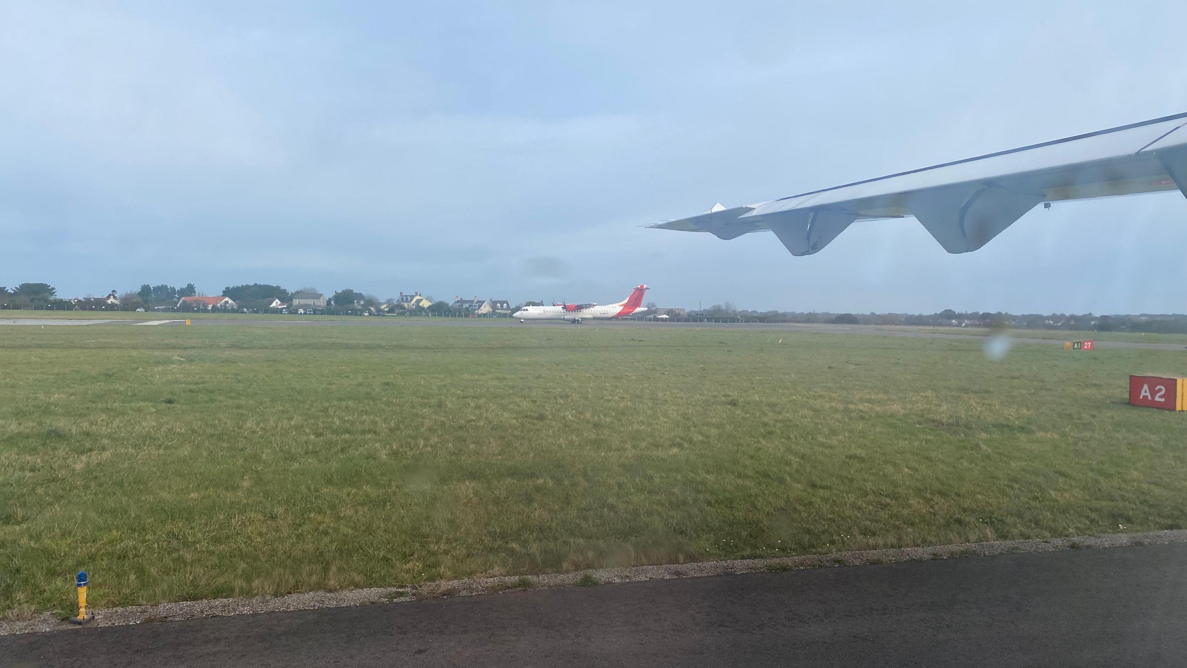 A grassy area on a airport runway. There is a plane in the distance which is white and red, the grass is green and there is a wing of a plane in the top right of the photo.