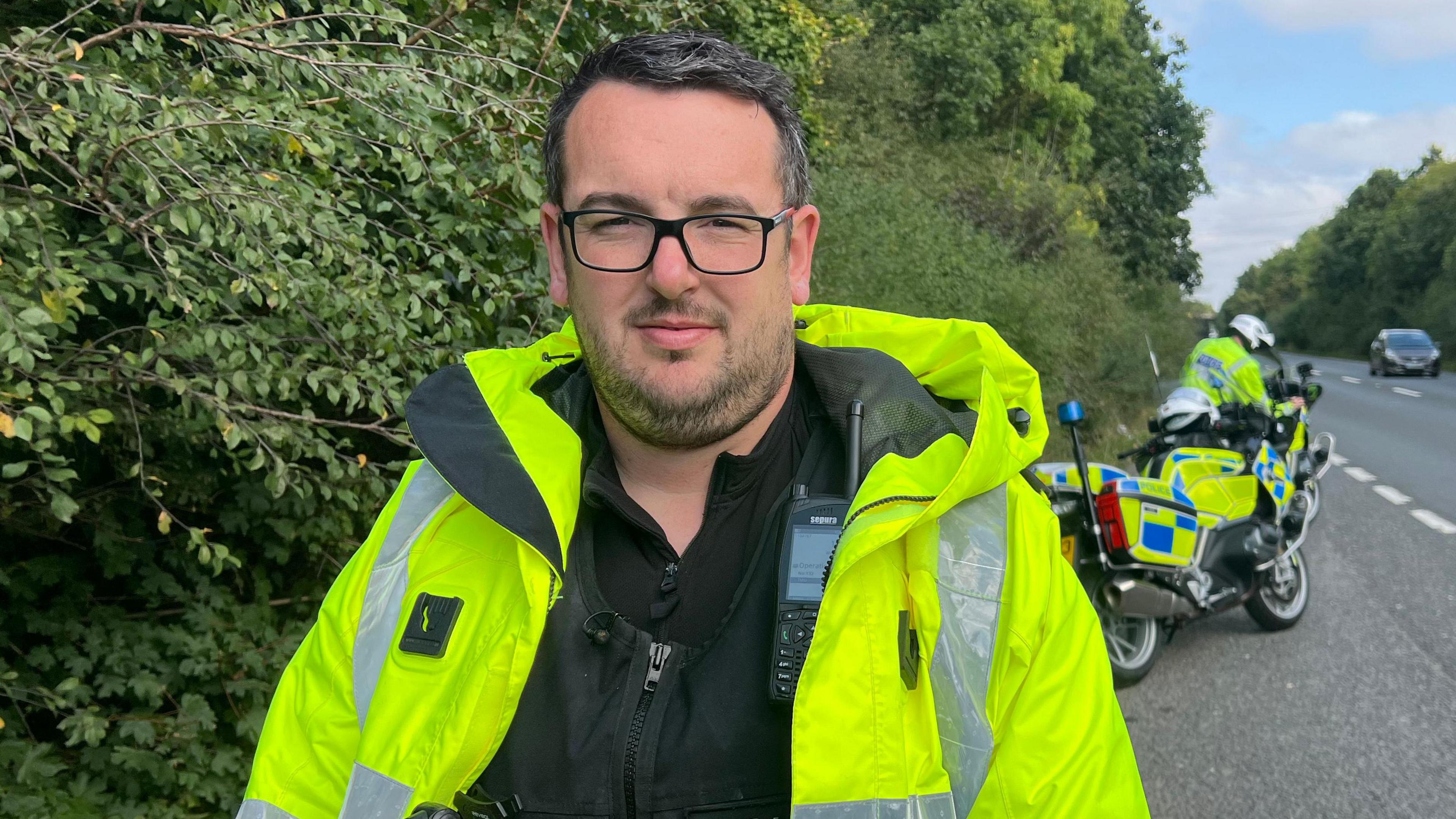 Matt Boiles wearing glasses and a hi-vis yellow coat over his police uniform, with a radio strapped to his chest. He is standing on the side of the road, with a police motorbike parked in the background.