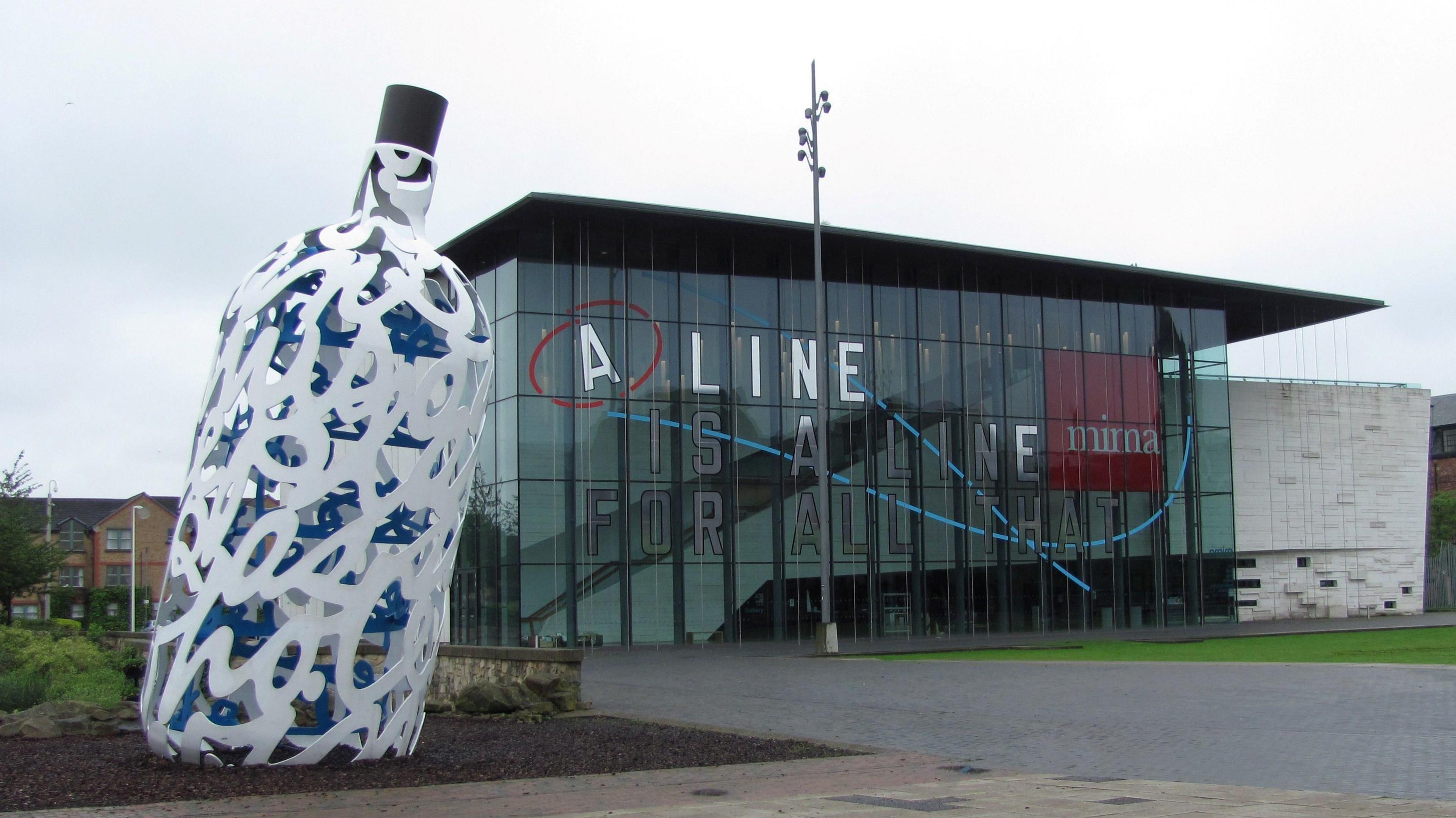 A general view of Mima in Middlesbrough. The building is made of glass and has a flat roof. There is a large sculpture of a white and blue bottle to one side.
