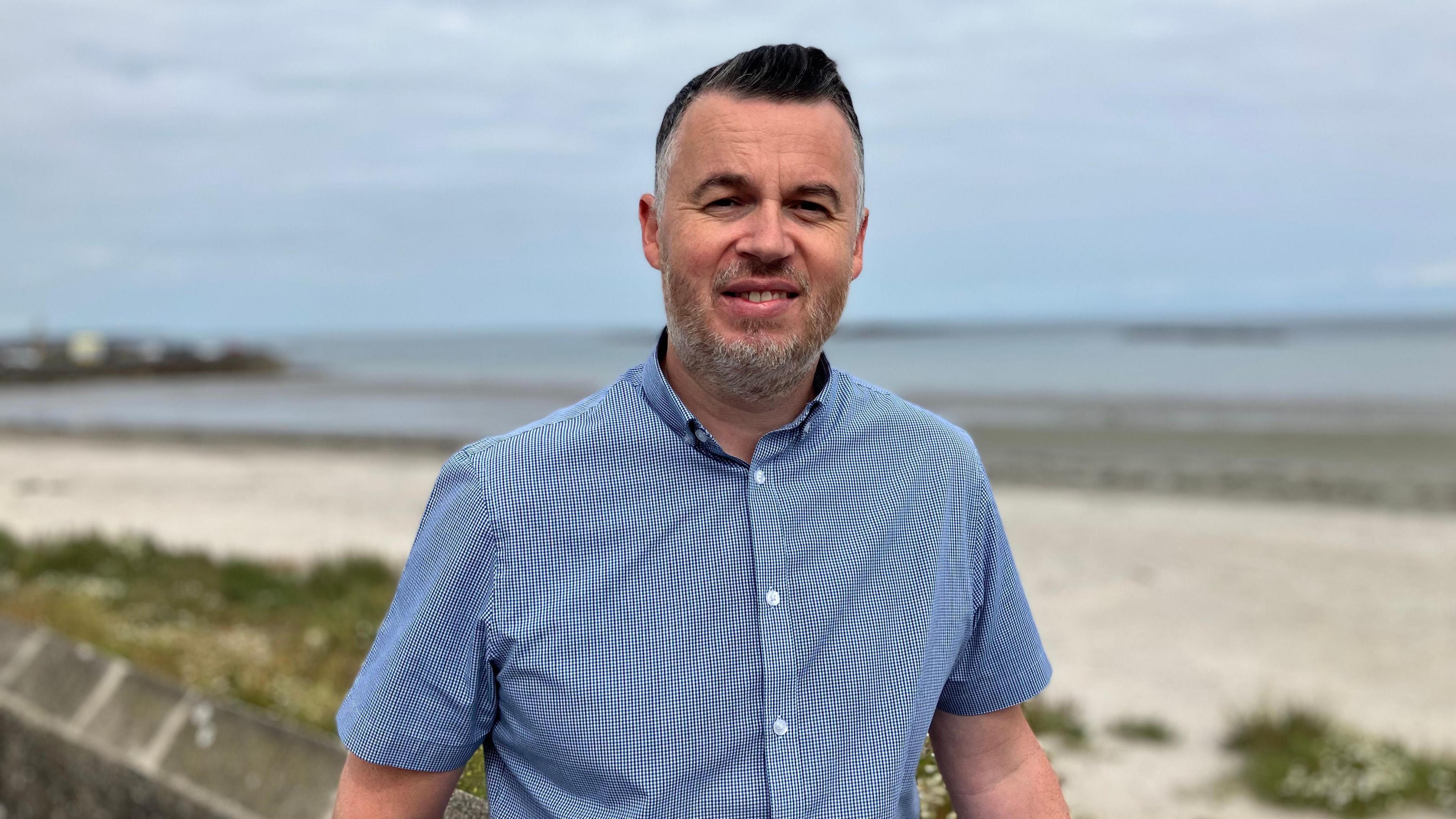 A man with short black hair and grey stubble, wearing a short sleeved blue shirt. He is standing in front of Millisle Beach.