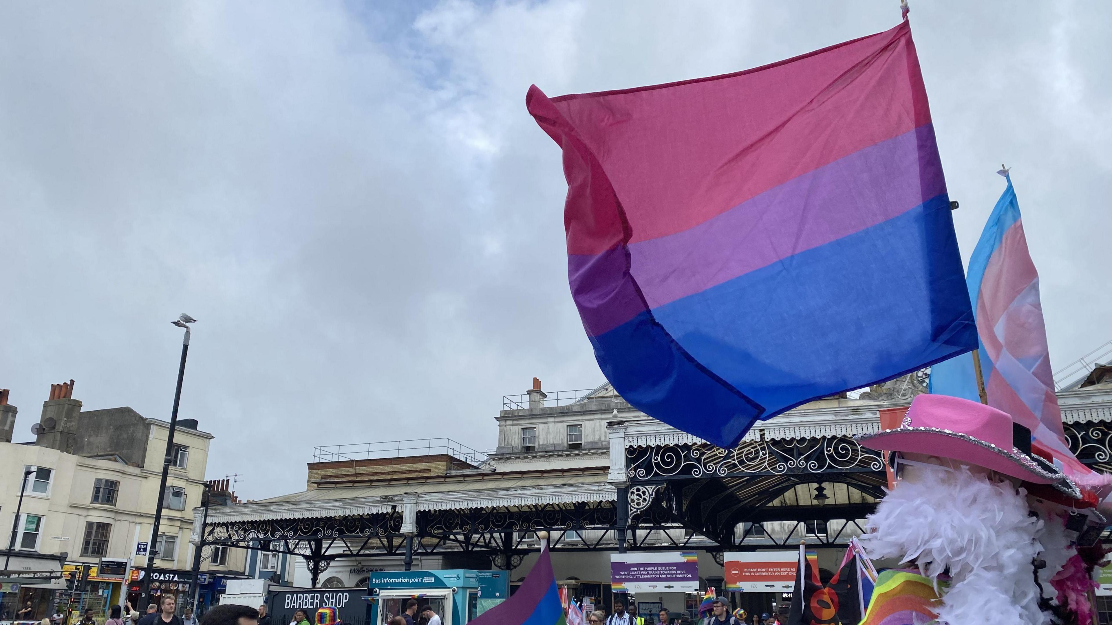 People selling rainbow flags outside Brighton Railway Station. The sky is most grey and cloudy, with a patch of blue