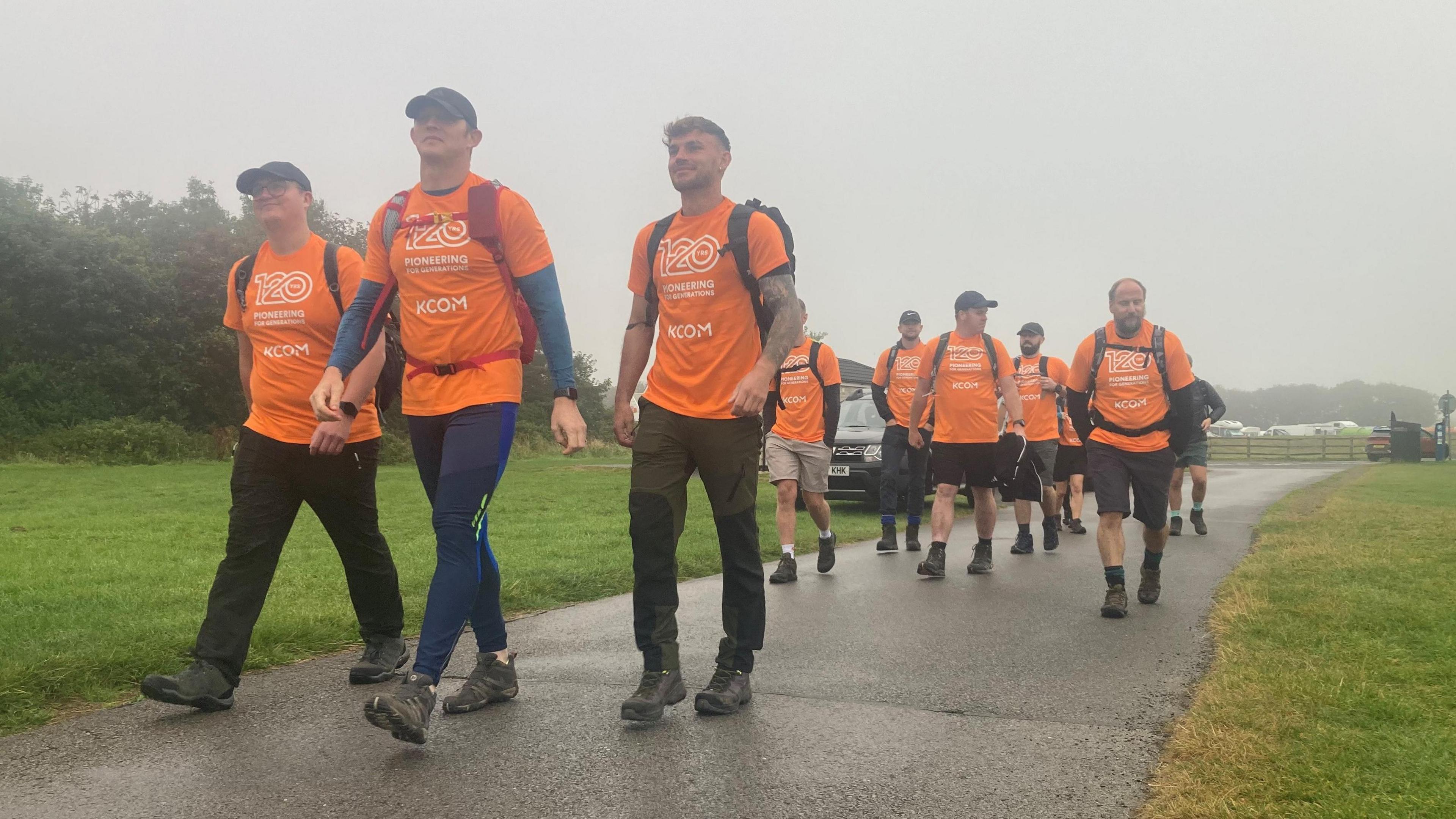 A group of hikers in matching orange T-shirts walk up a hill towards the camera.