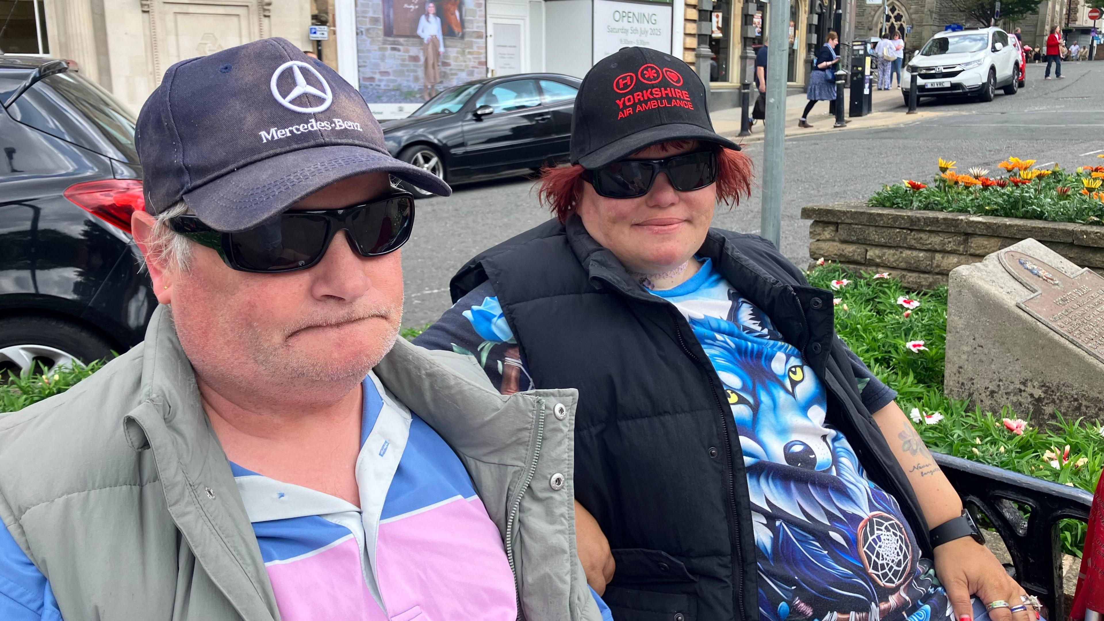 Paul Flatters is on the left of the picture, his wife, Rachel, is on the right. They are both wearing baseball caps and sunglasses. Rachel's T-shirt has an image of a blue wolf with yellow eyes.