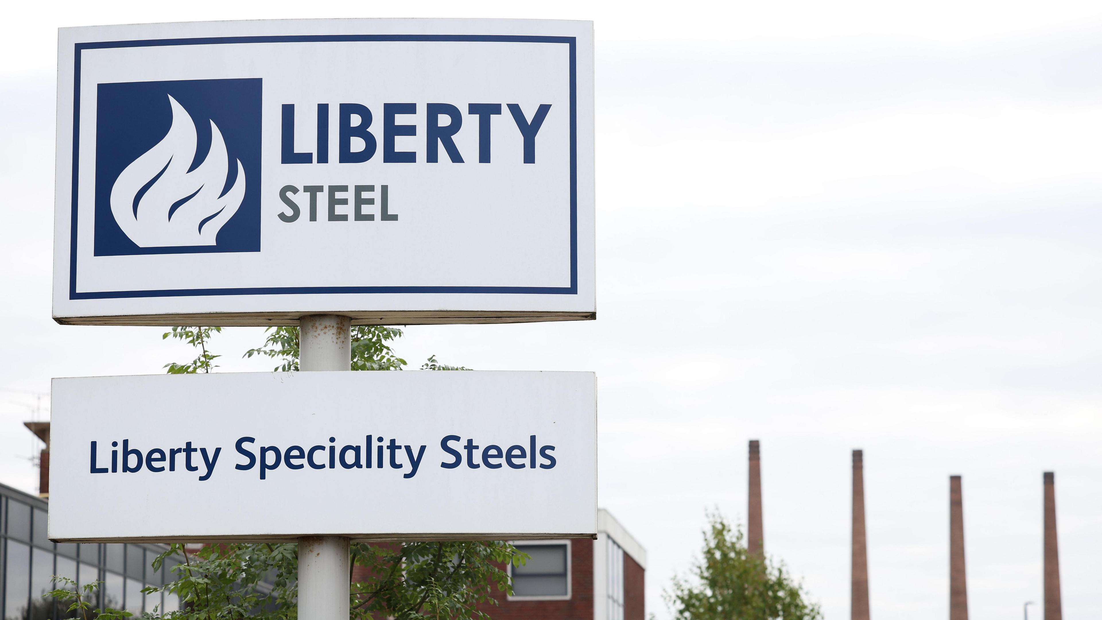 A white sign bearing the words 'Liberty Steel' and 'Liberty Speciality Steels' is in the foreground to the left. In the background to the right, four tall steelworks chimneys can be seen, along with a cloudy sky and the tops of trees.