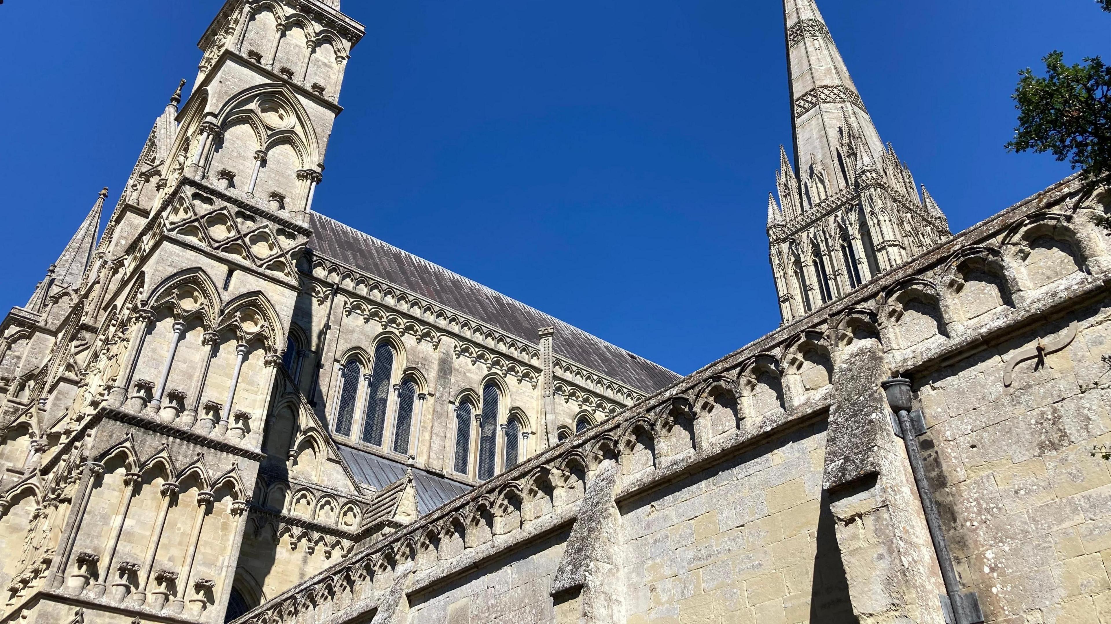 A partial view of Salisbury Cathedral taken from outside the cloister, looking up towards the spire, set against a royal blue sky.
