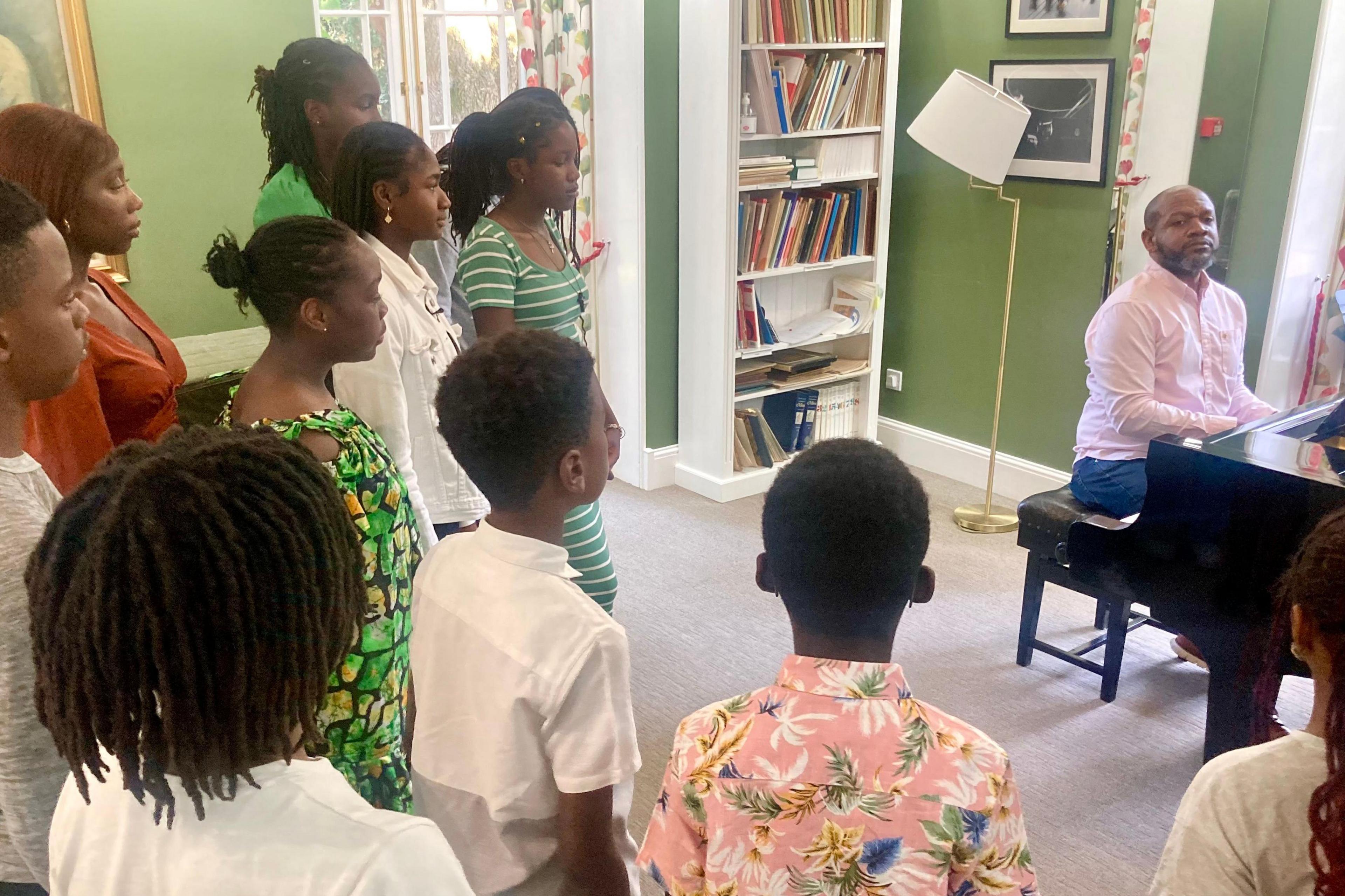 Dr Jeffrey Allen Murdock sits at a piano, wearing a pink shirt, as children wearing brightly coloured clothes stand around him, ready to sing for choir practice.