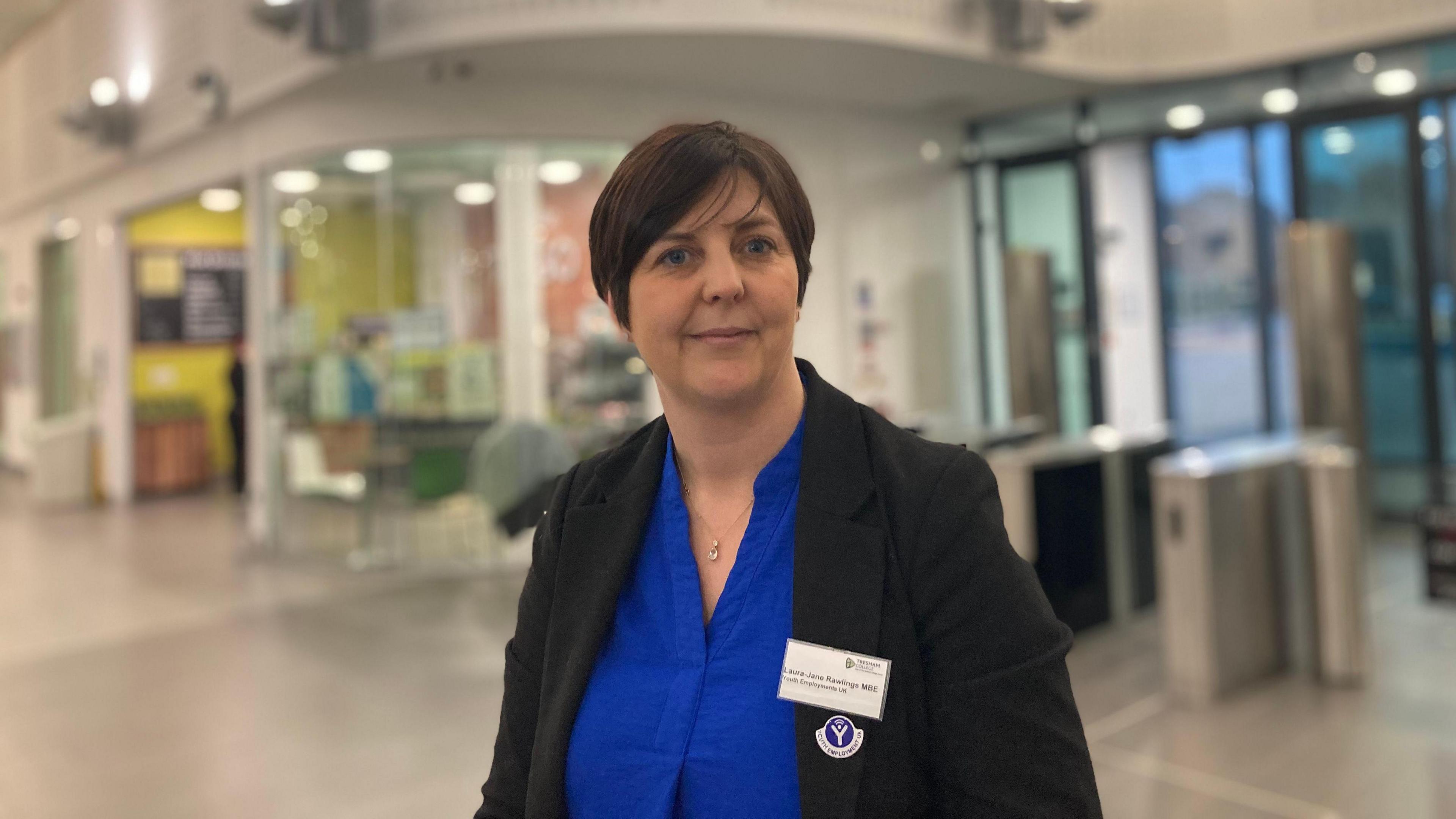 A woman in a blue shirt and black blazer stands in the centre of a corridor looking at the camera. 