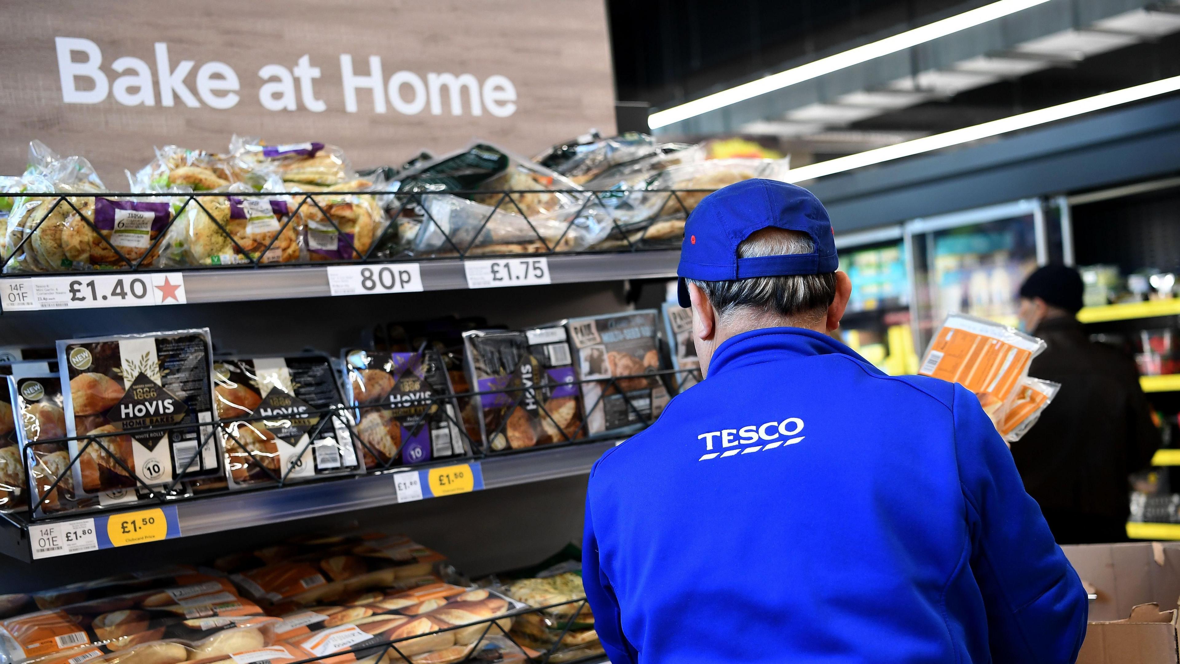 A man wearing blue Tesco uniform is restocking a shelf in the bakery section