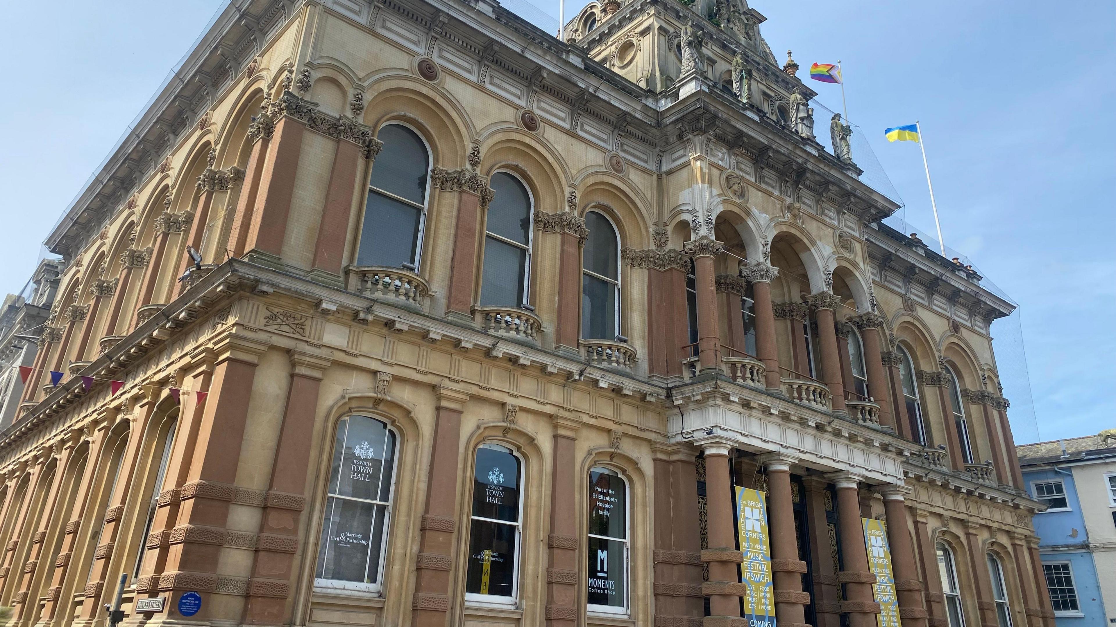 The exterior and entrance to Ipswich Borough Council's town hall. It is a palacial building with pillars and statues. The entrance to the council is accompanied by posters hanging up from the columns and flags waving on the roof.