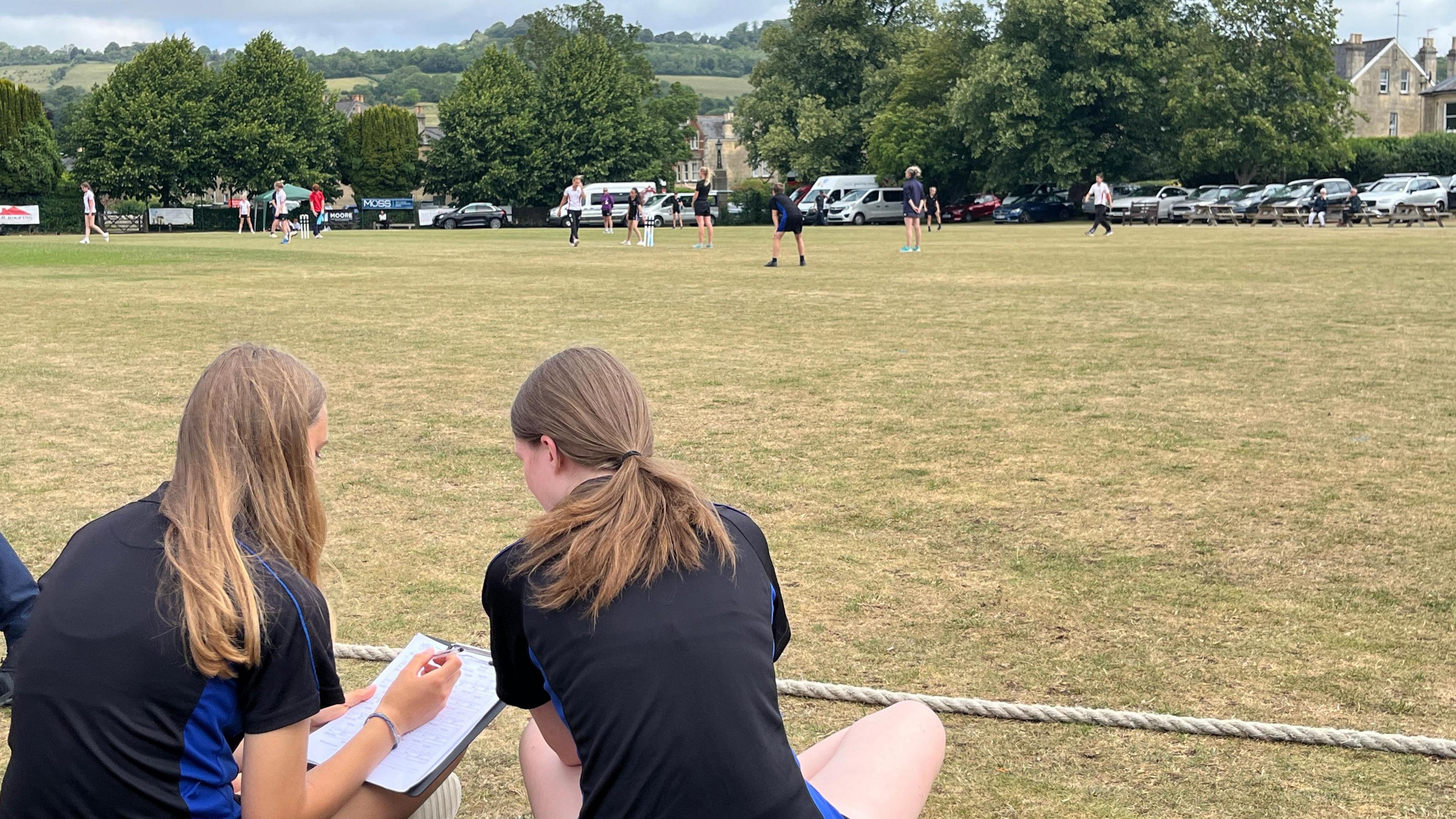 Two girls sit on the boundary of the Lansdown Cricket club pitch. They are watching a match taking place in the distance. The weather is dry and the cricket pitch is yellowed and hard.