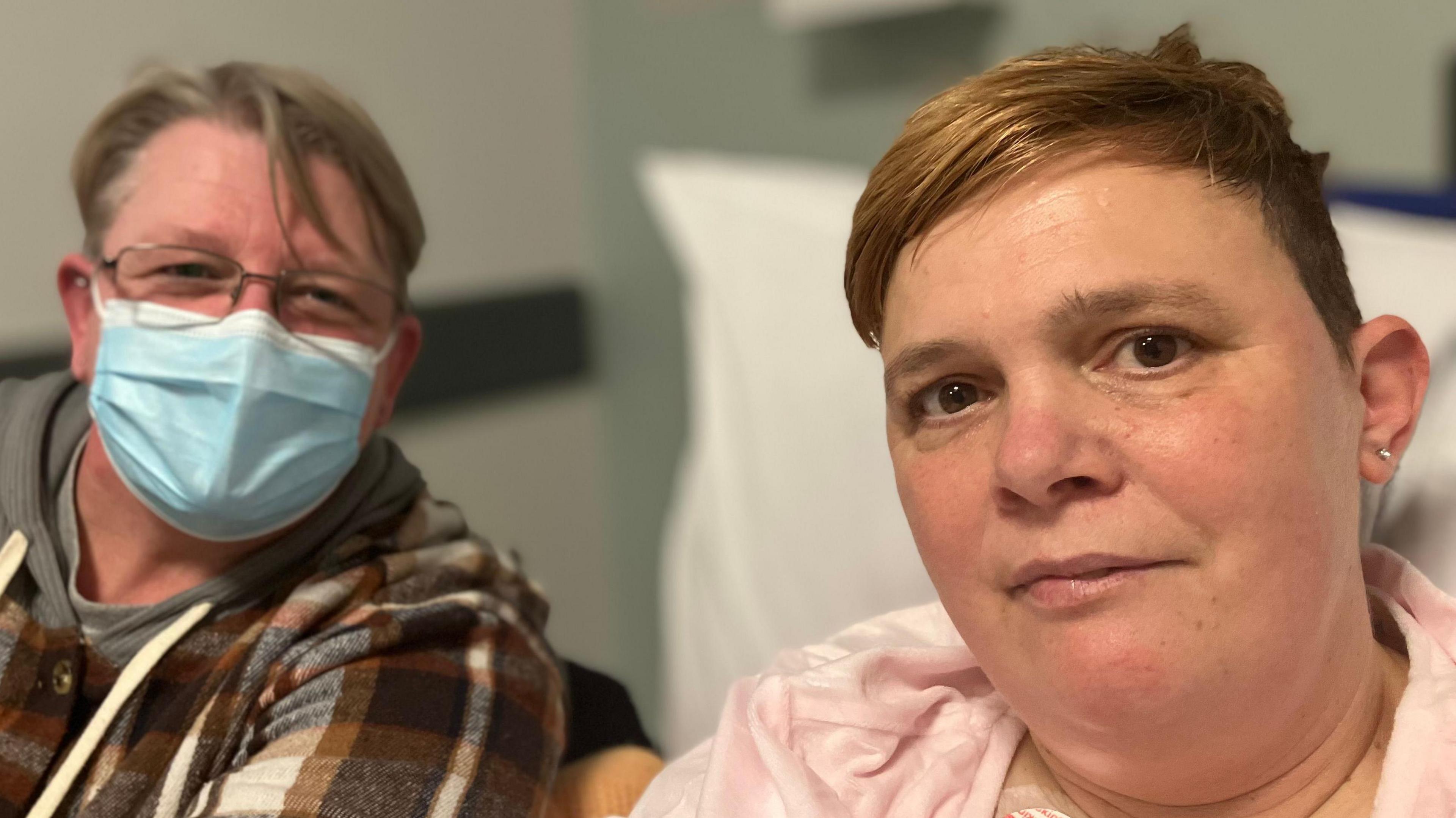 Natalie and her partner Karen sitting in the emergency department. Karen Davies (left) has short fair hair and is wearing a chequered brown and cream jacket and a blue face mask. Karen has short brown hair and is wearing a pink panama top.