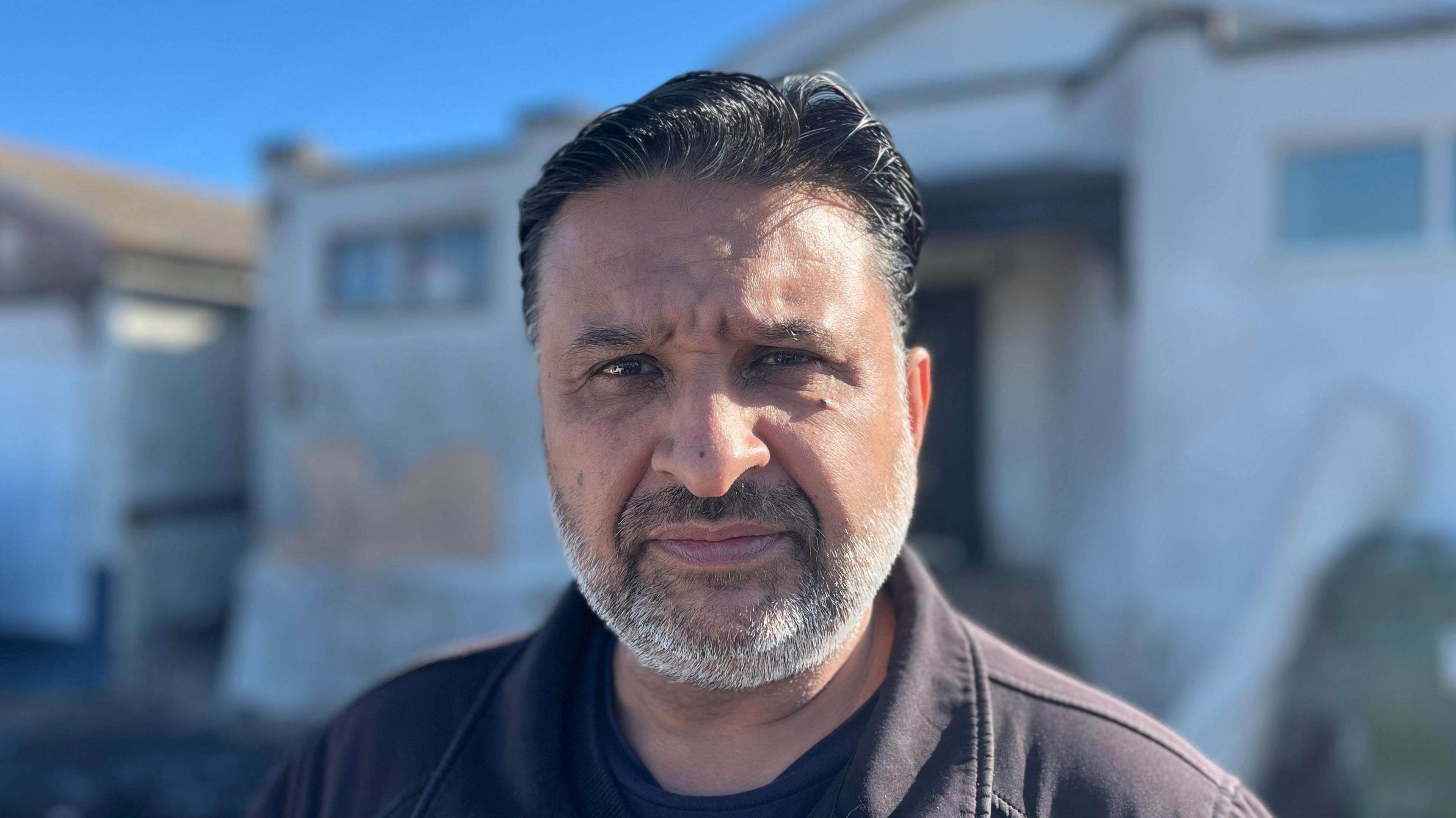 A man with short black hair and grey stubble. He is wearing a black jacket and is looking into the camera while stood in front of a white building.