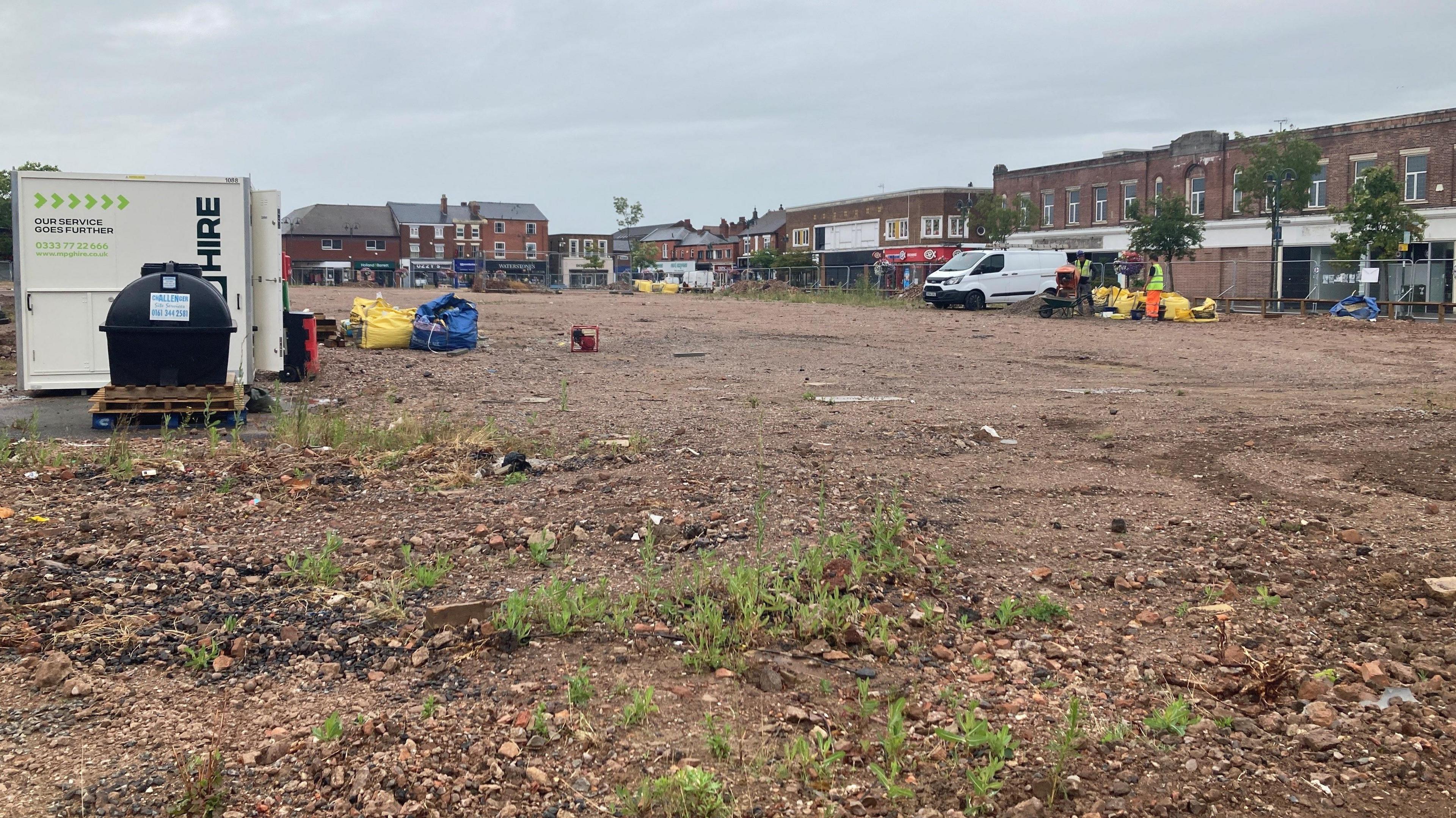 A view of the empty Royal Arcade site. It is a large swathe of dry, exposed soil with a few tufts of grass. There are shops and buildings around the permiter. There is a white van and some equipment on site, including a metal containers and some large materials bags.