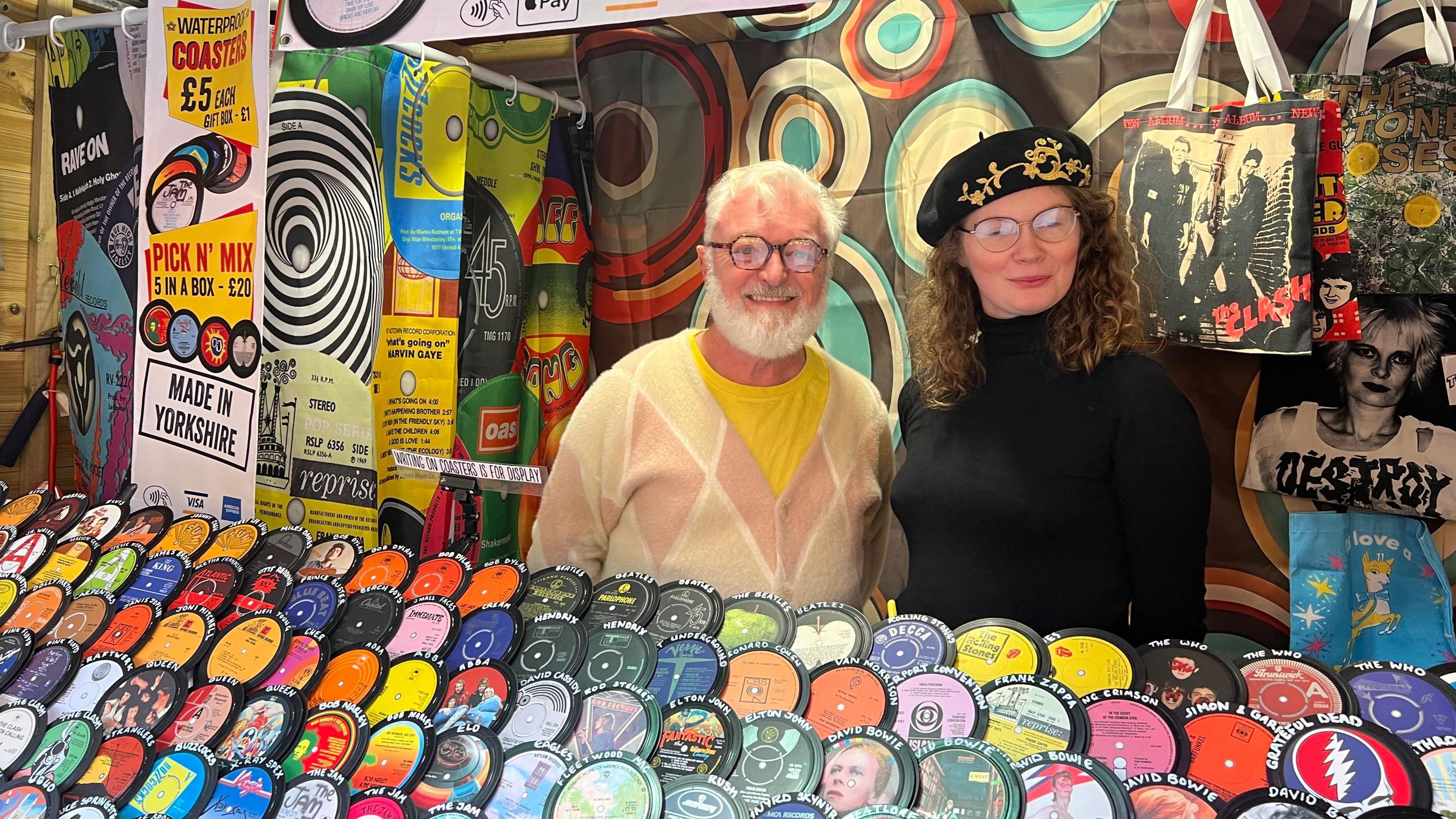 Two people smiling next to an array of record label coasters. The man on the left has glasses, grey hair and a grey beard and is smiling. He is stood next to a smiling woman with curly brown hair, who is wearing glasses, a black beret and a black turtleneck top.