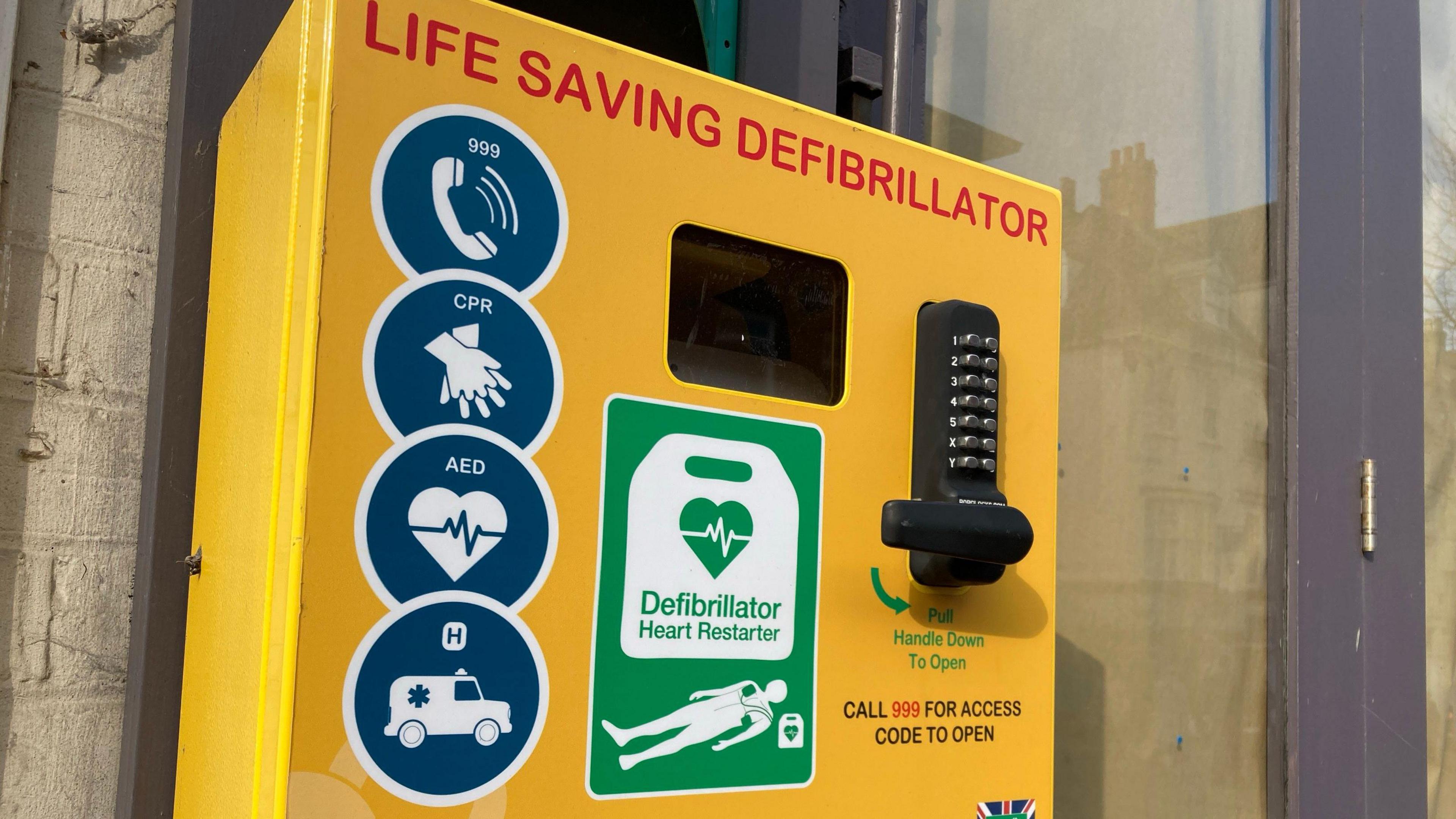 A defibrillator in a yellow box attached to a wall