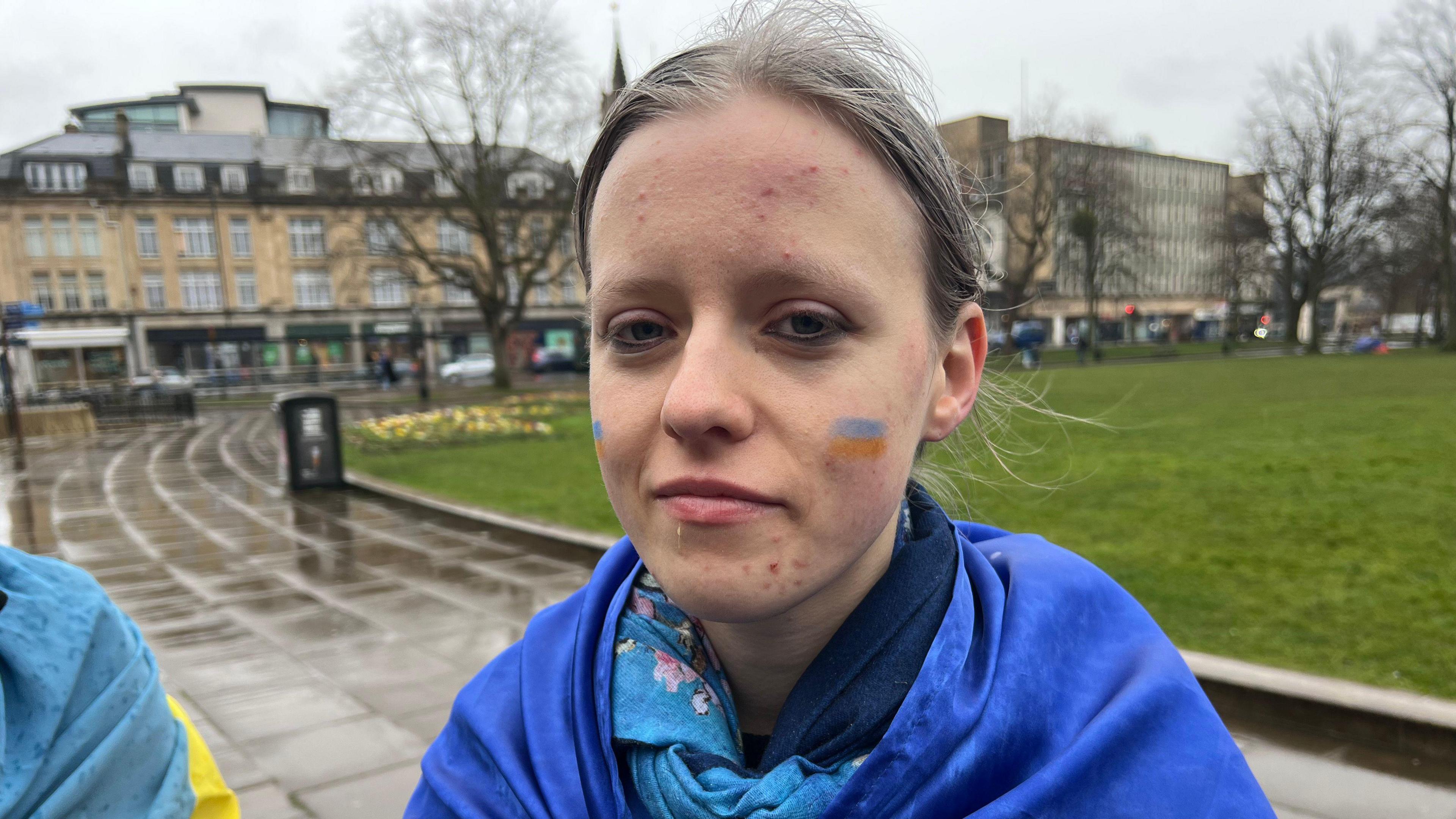 Yuliia Shynkarenko standing beside College Green on a wet and grey day. She is wrapped in a blue and yellow Ukranian flag, with two small Ukranian flags painted on each of her cheeks. Her hair is pulled back into a ponytail and she is wearing black eyeliner. She is looking at the camera with a sad but determined expression.