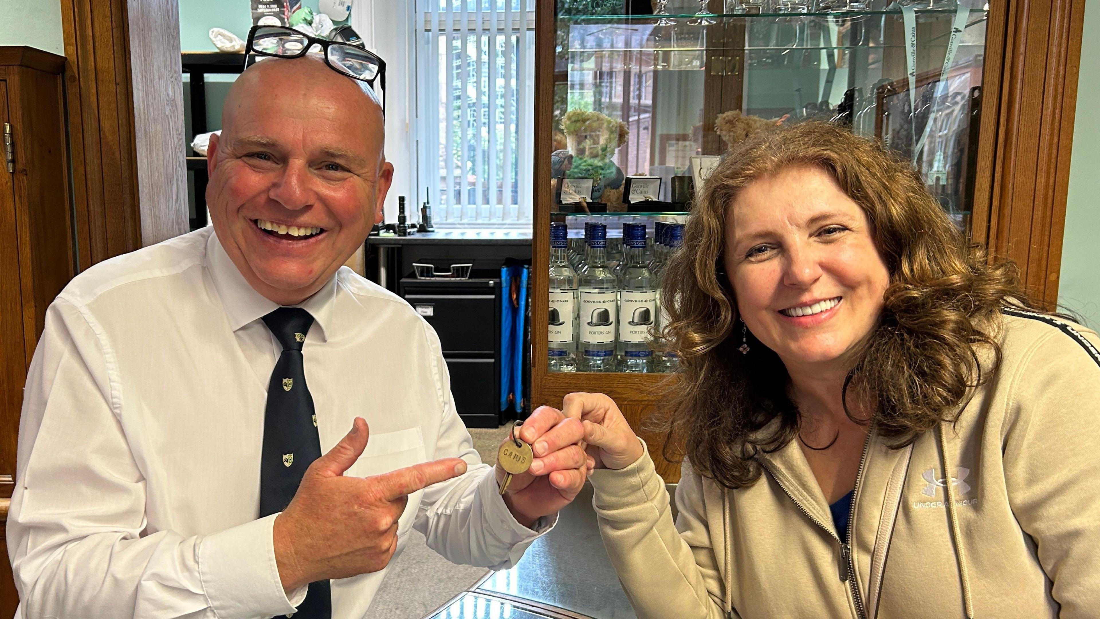 Senior porter John Turton is on the left of the image and has spectacles balanced on the top of his balding head. He is wearing a white shirt and a black tie with college emblems on it. Carla Risoldi, on the right has wavy shoulder length brown hair and is wearing a light brown jacket. Mr Turton is holding a small key between them and he is pointing at it.