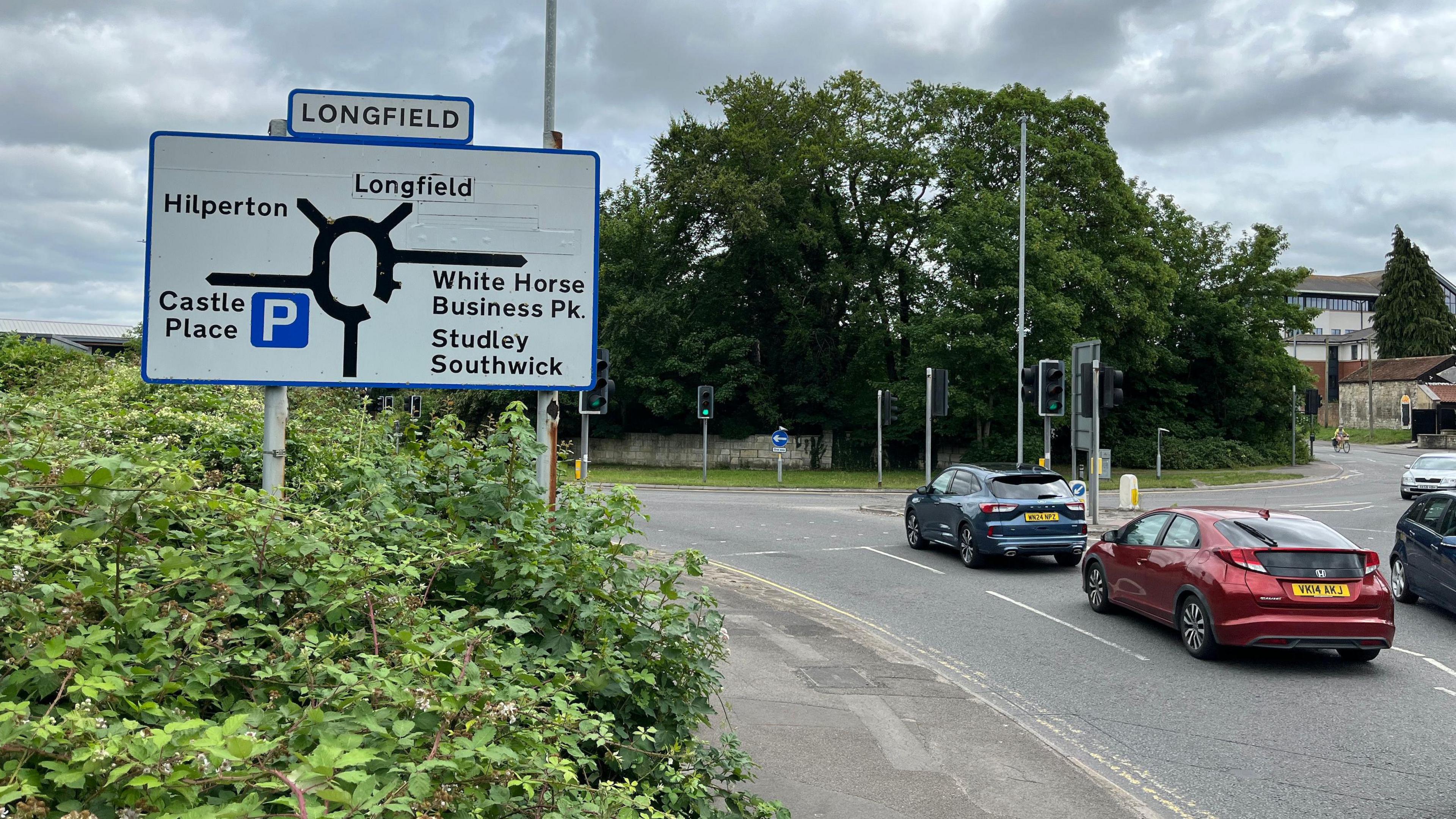 A large road sign showing a gyratory system with roads leading to Longfield, Hilperton, Castle Place, White Horse Business Park and Studley. Cars are driving past the sign.