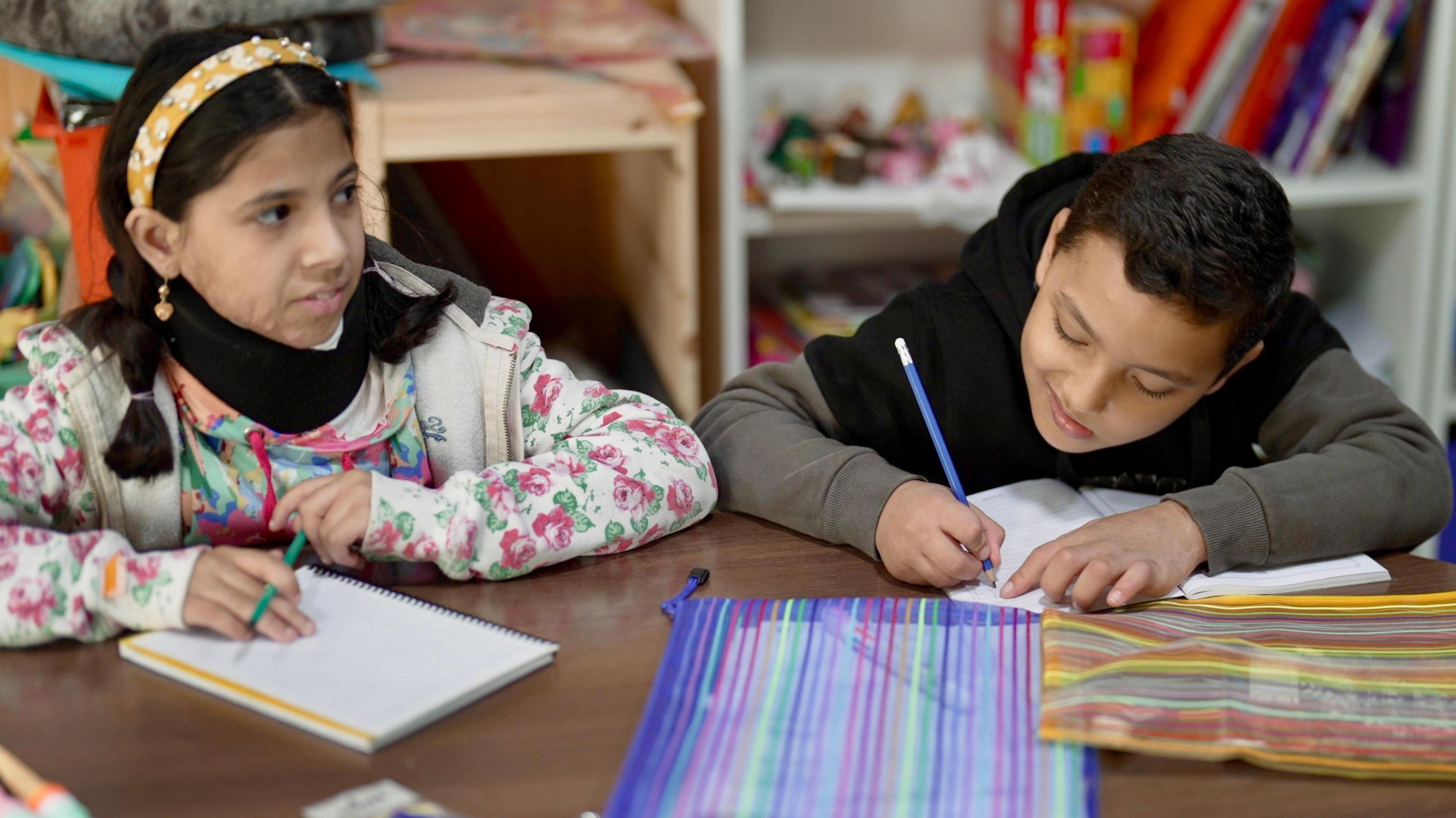 A girl and a boy sit at a table writing with colourful pencil cases in front of them