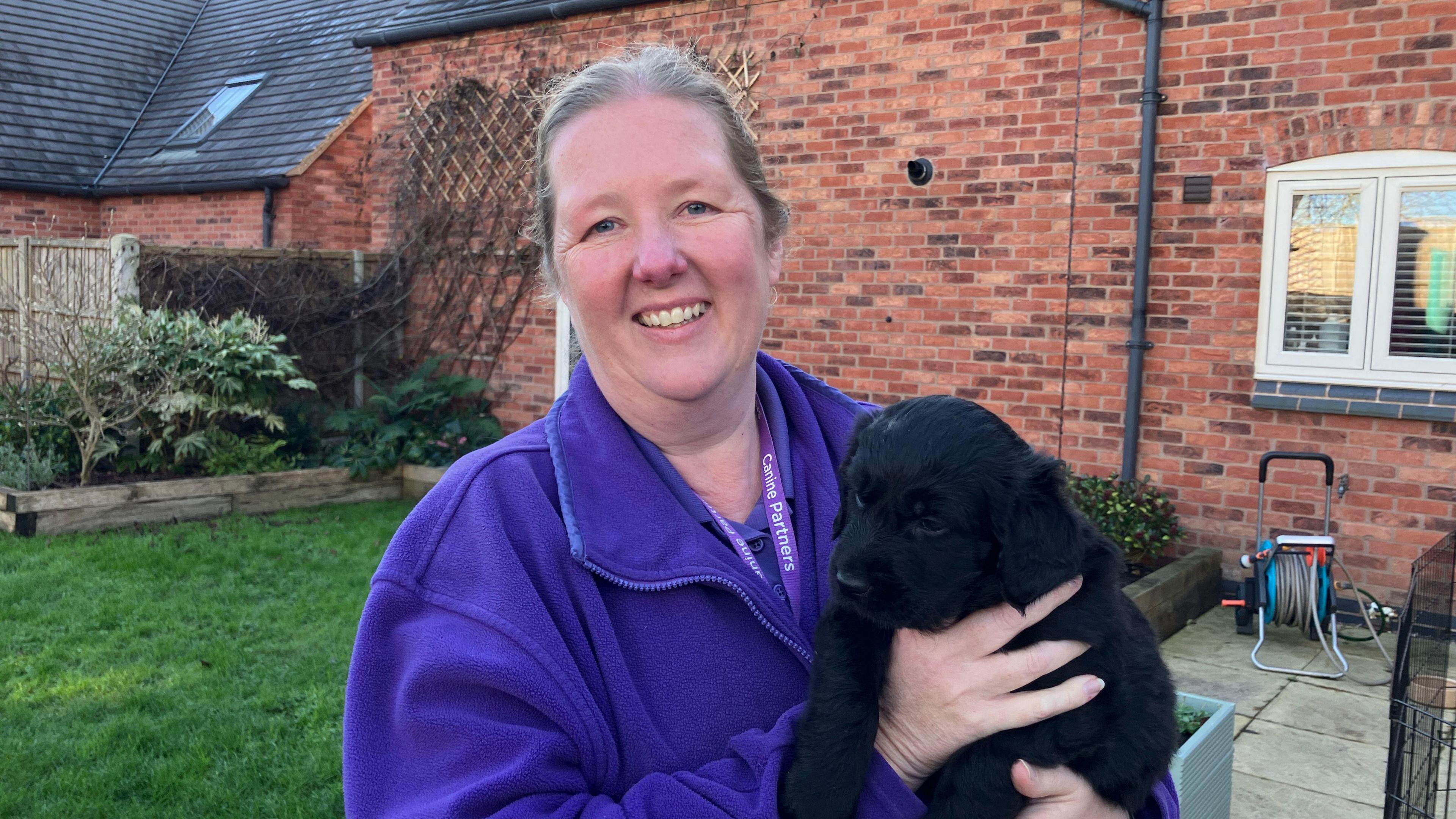 A woman with grey hair tied back. She is wearing a dark purple jumper. She is holding a black labradoodle puppy. Behind her is a green lawn and a patio as well as a house