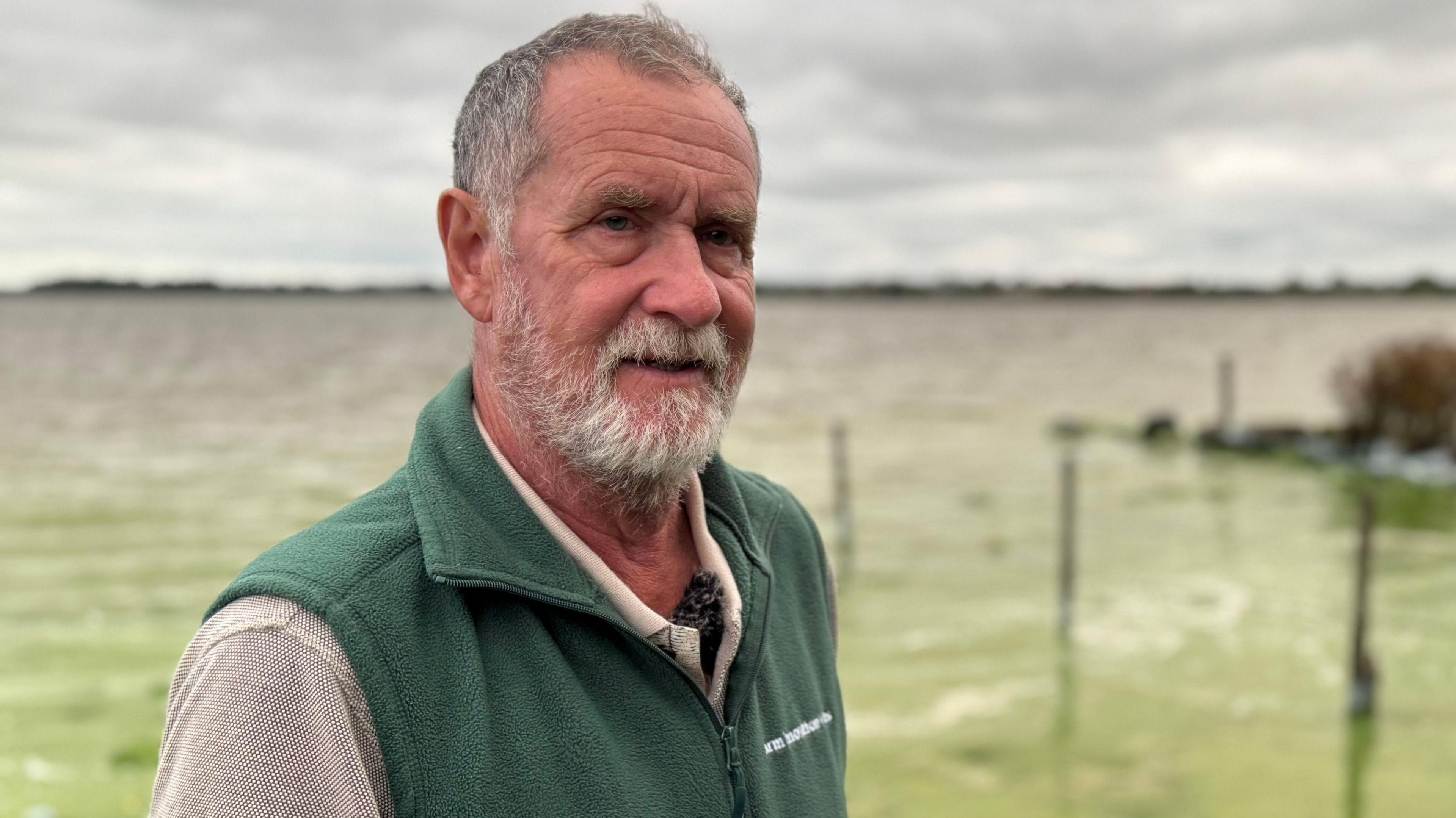 Michael Meharg is an older man with short grey hair and a white beard. He is standing in front of the algae on Lough Neagh but it is blurred. He is wearing a grey shirt and a green gillet fleece.