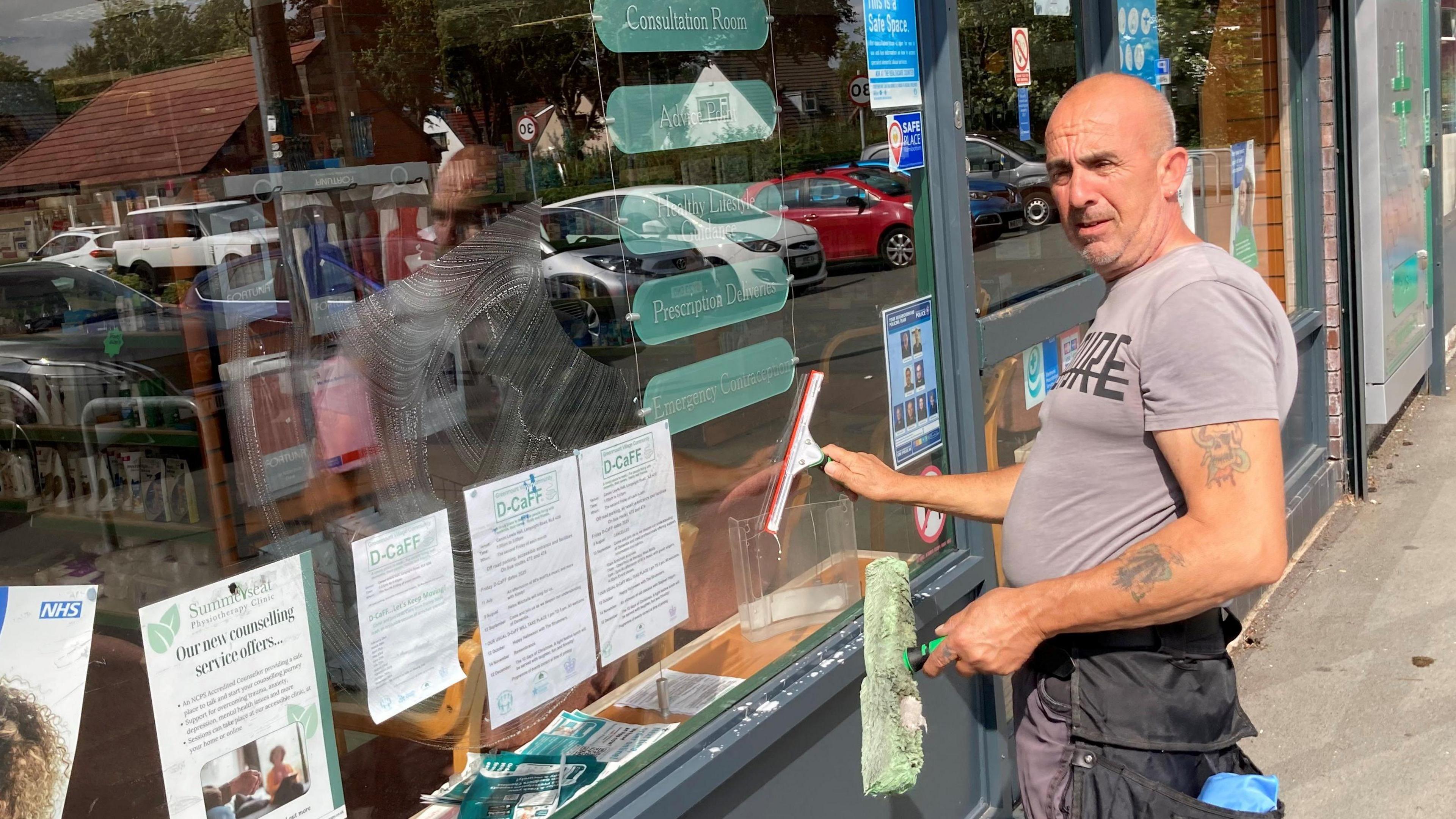 Raymond Pavitt is standing in front of a pharmacy window with his window cleaning equipment. He is wearing a grey T-shirt