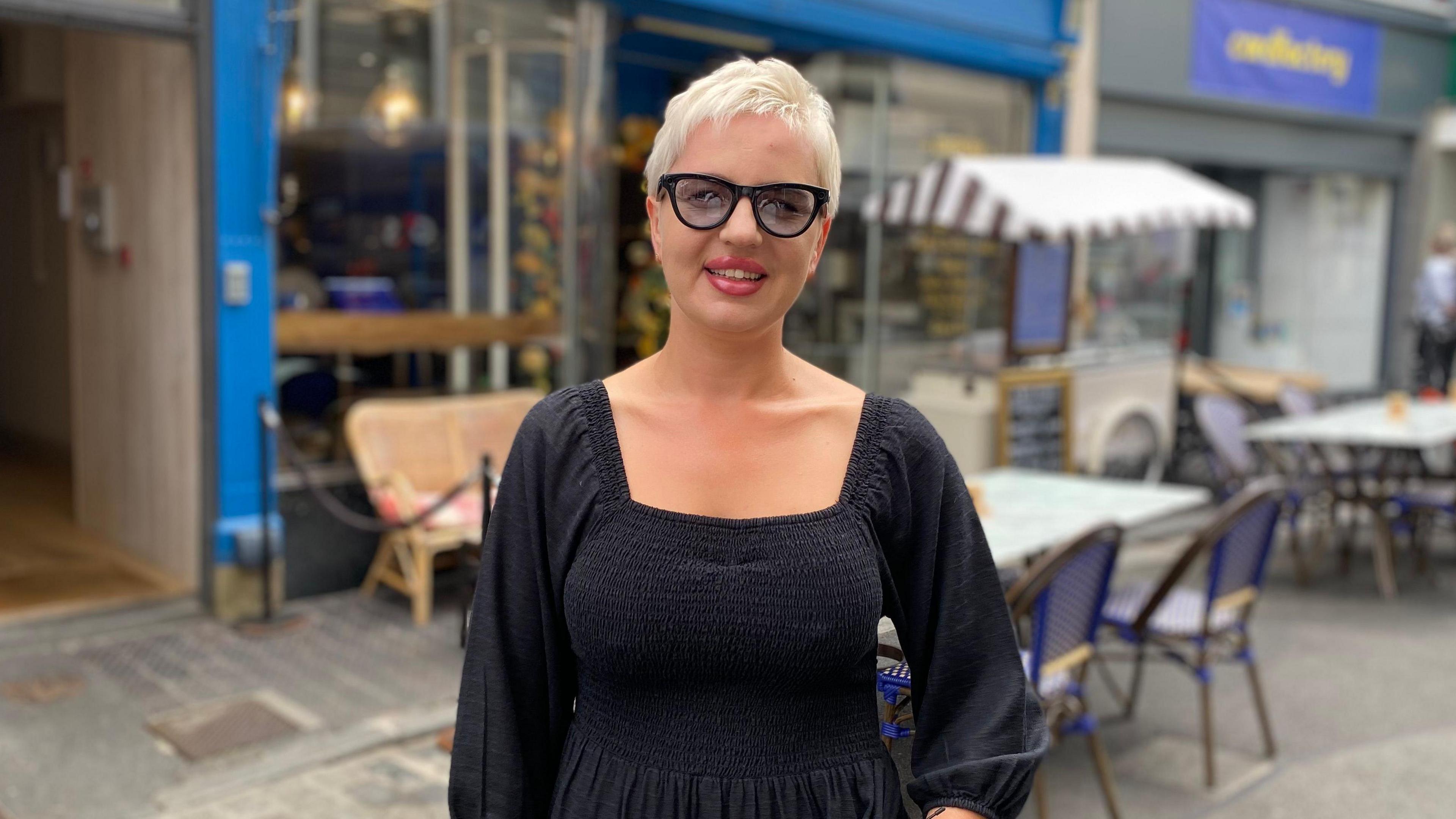 Natalia has short blonde hair. She is wearing black framed glasses and a black dress.