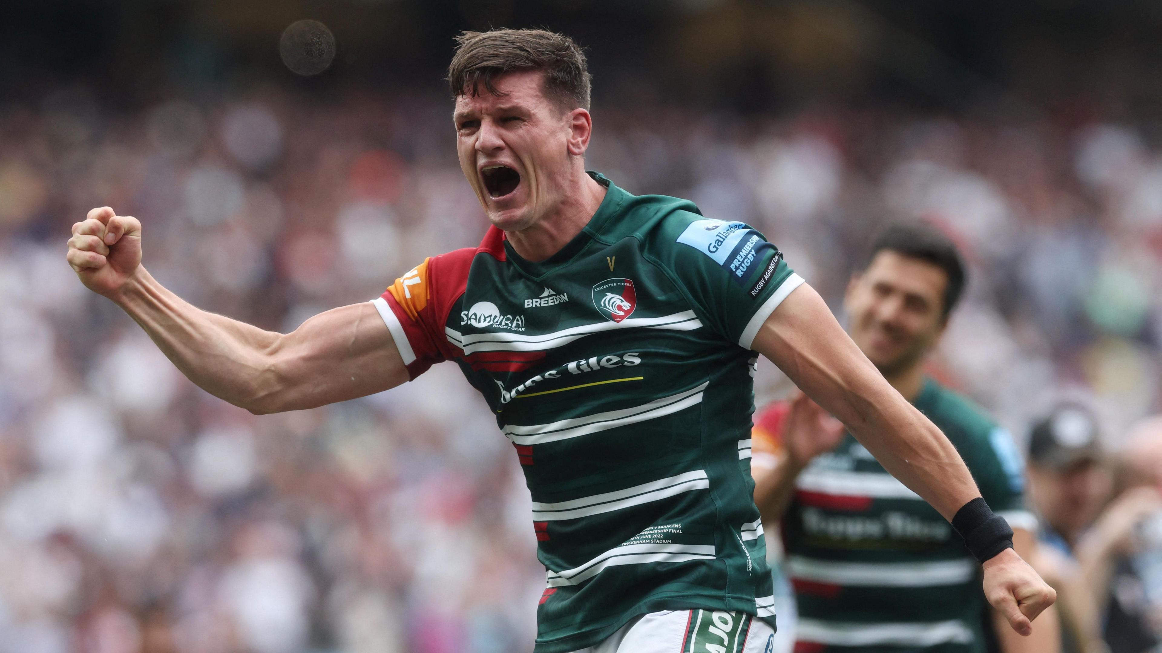 Freddie Burns holds his right arm out with his fist clenched in celebration after Leicester win the Premiership title in 2021. He is standing on the pitch at Twickenham wearing green and white striped Leicester kit