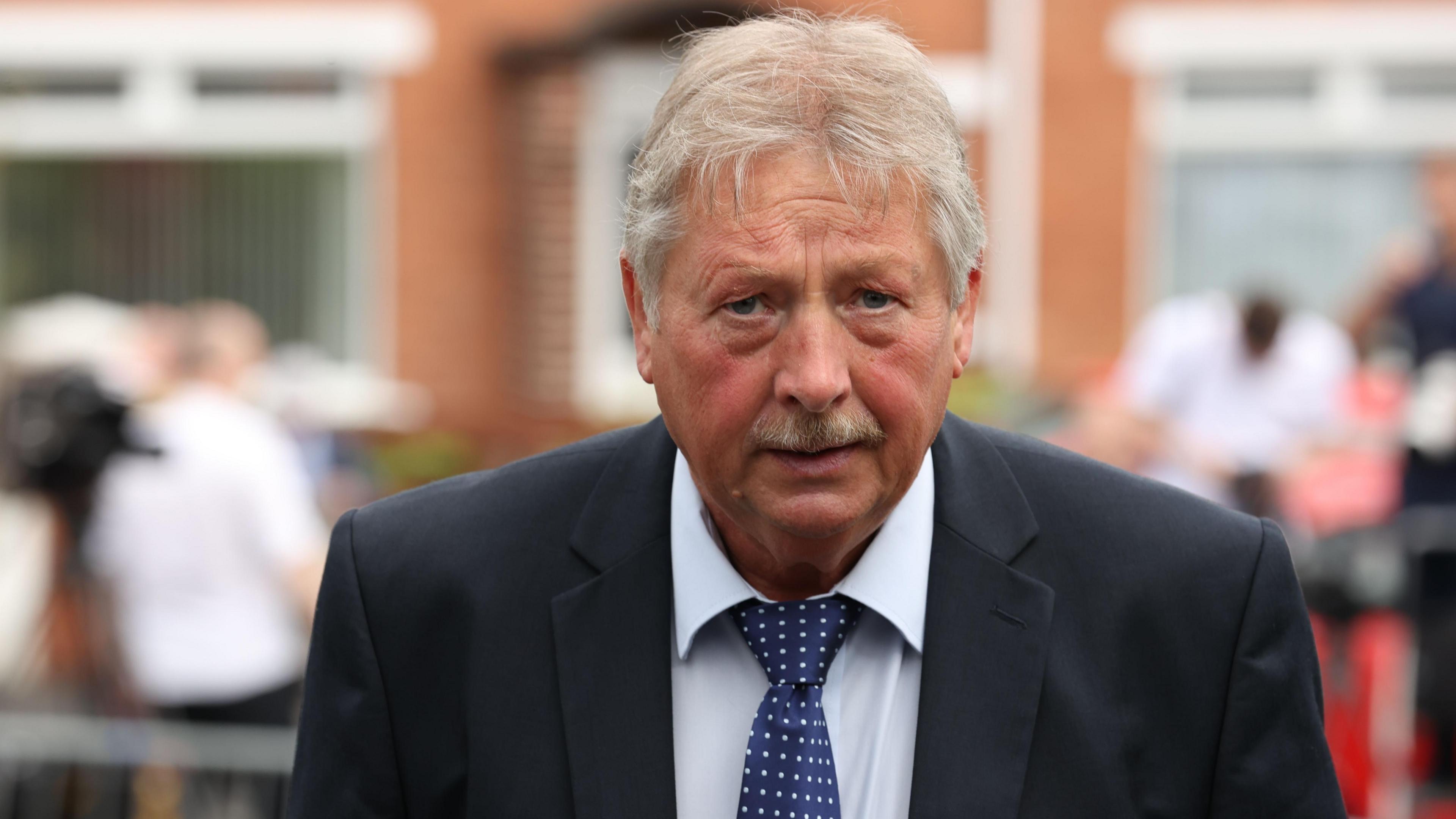 Sammy Wilson looking at the camera with a neutral expression. He is wearing a suit and tie. There are people and a building in the background very out of focus.