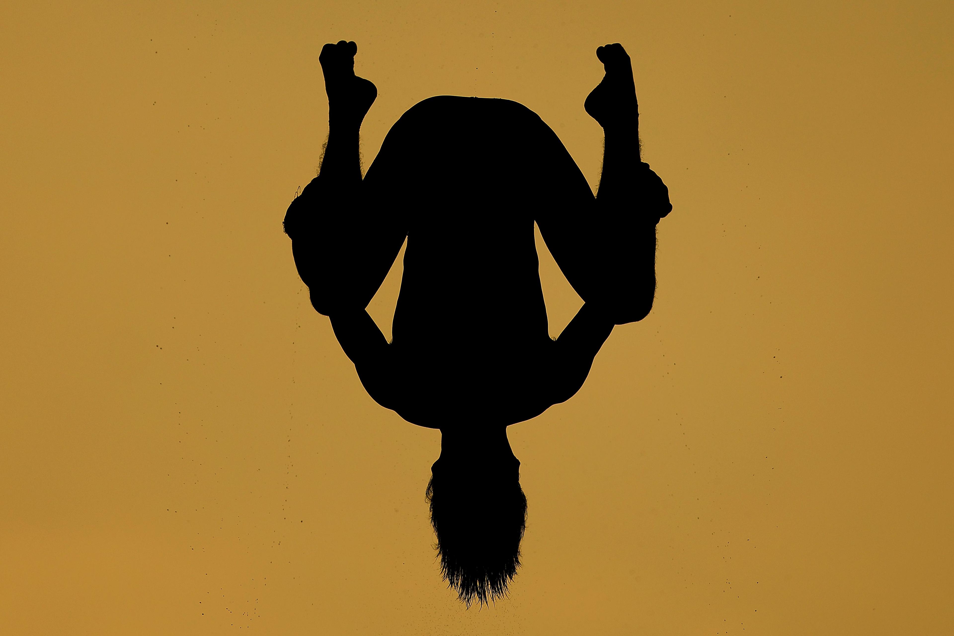 A silhouetted diver is captured mid-air against a golden background, body inverted with knees bent and arms gripping the legs. Water droplets scatter around the figure, emphasizing motion and the dramatic contrast between the dark silhouette and bright backdrop.