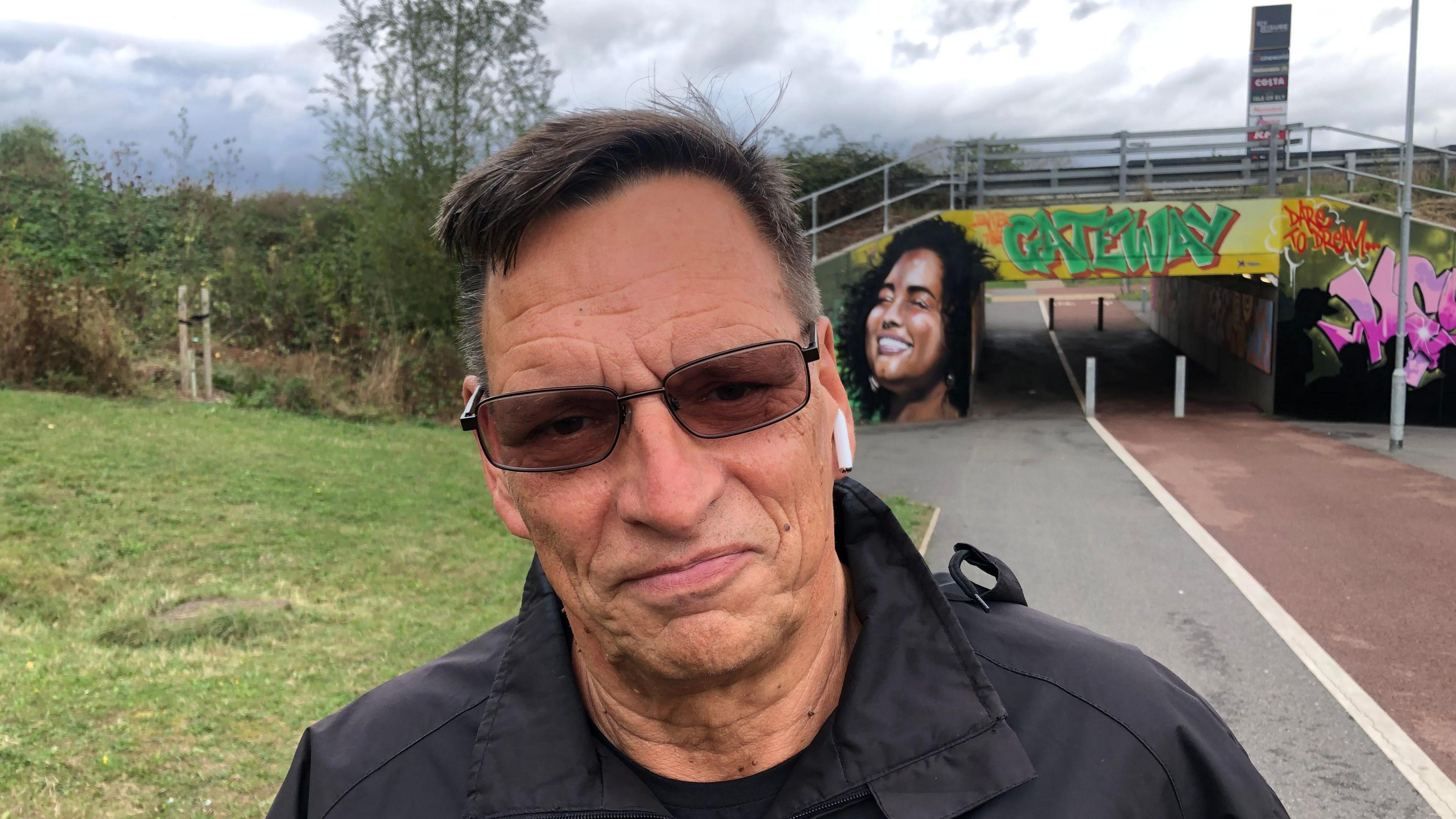 Steve Kendall, 64, a man with black short hair, sunglasses and ear pods standing in front of the colourful underpass beneath the A10 in Ely.
The pathway is red tarmac on one side for cycles and grey the other, for pedestrians.
A painting of a  dark haired smiling lady can be seen over the mans left shoulder