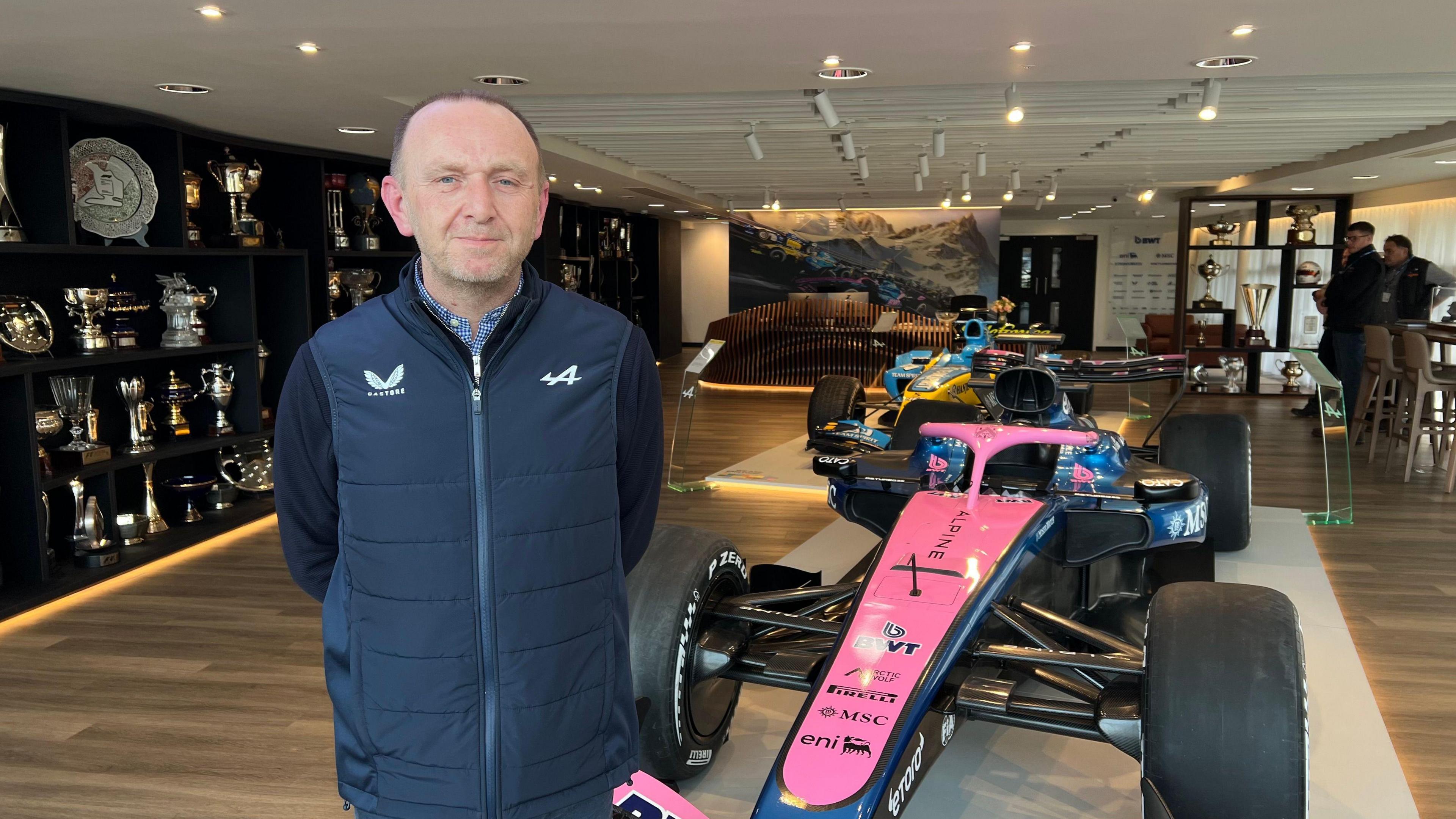 Ian Pearce, operations director at Enstone-based Alpine, posing for a picture in front of one of their cars. Trophies can be seen in a glass cabinets behind him. Two people can be seen looking at the cars in the far right corner of the room.