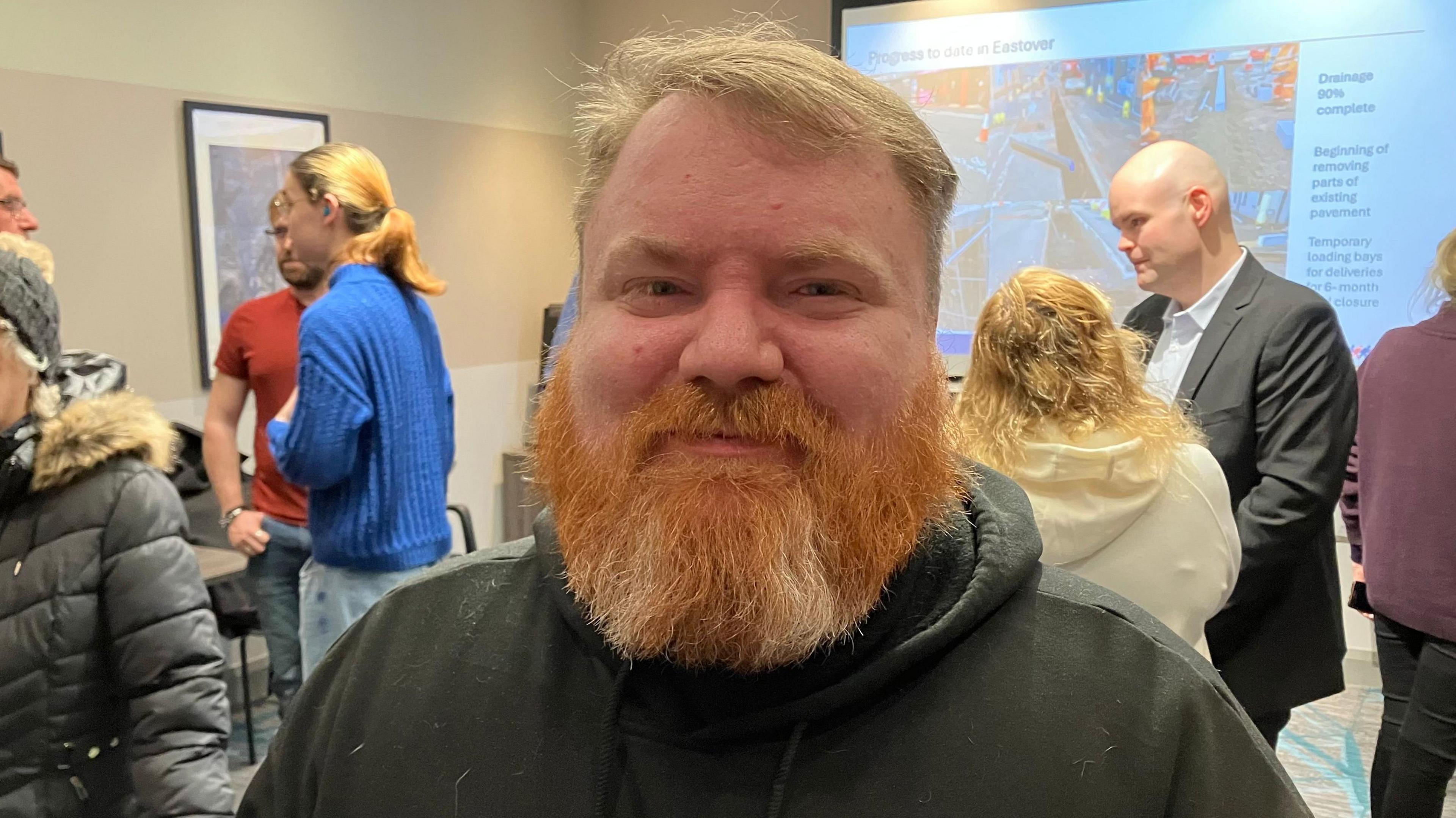 Man with ginger beard look at the camera with a few people behind him chatting to each other