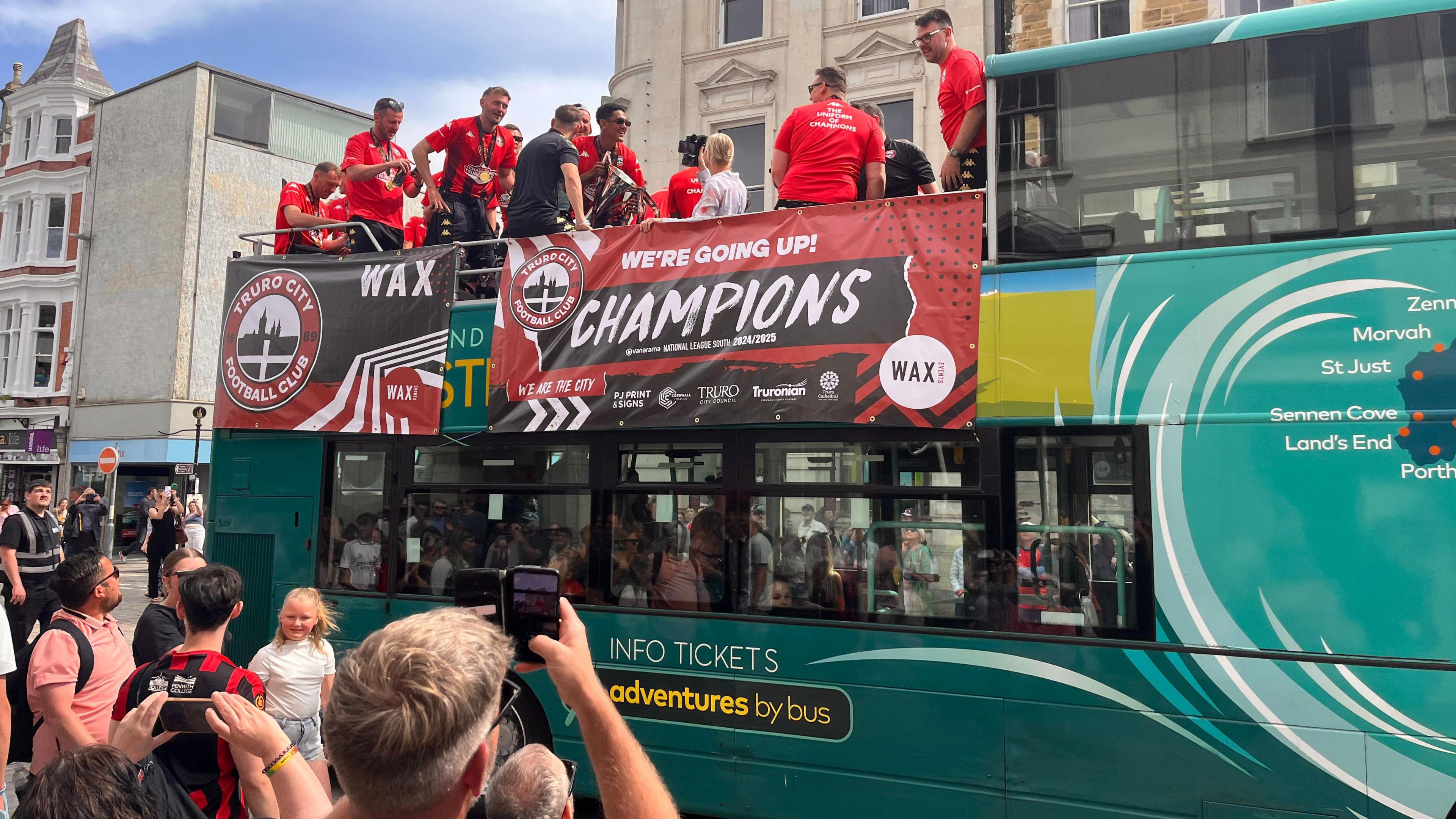 Players are on the top deck of the open bus and it is in the middle of the city. There are spectators looking up and taking photos. A banner on the bus reads: WE'RE GOING UP. CHAMPIONS