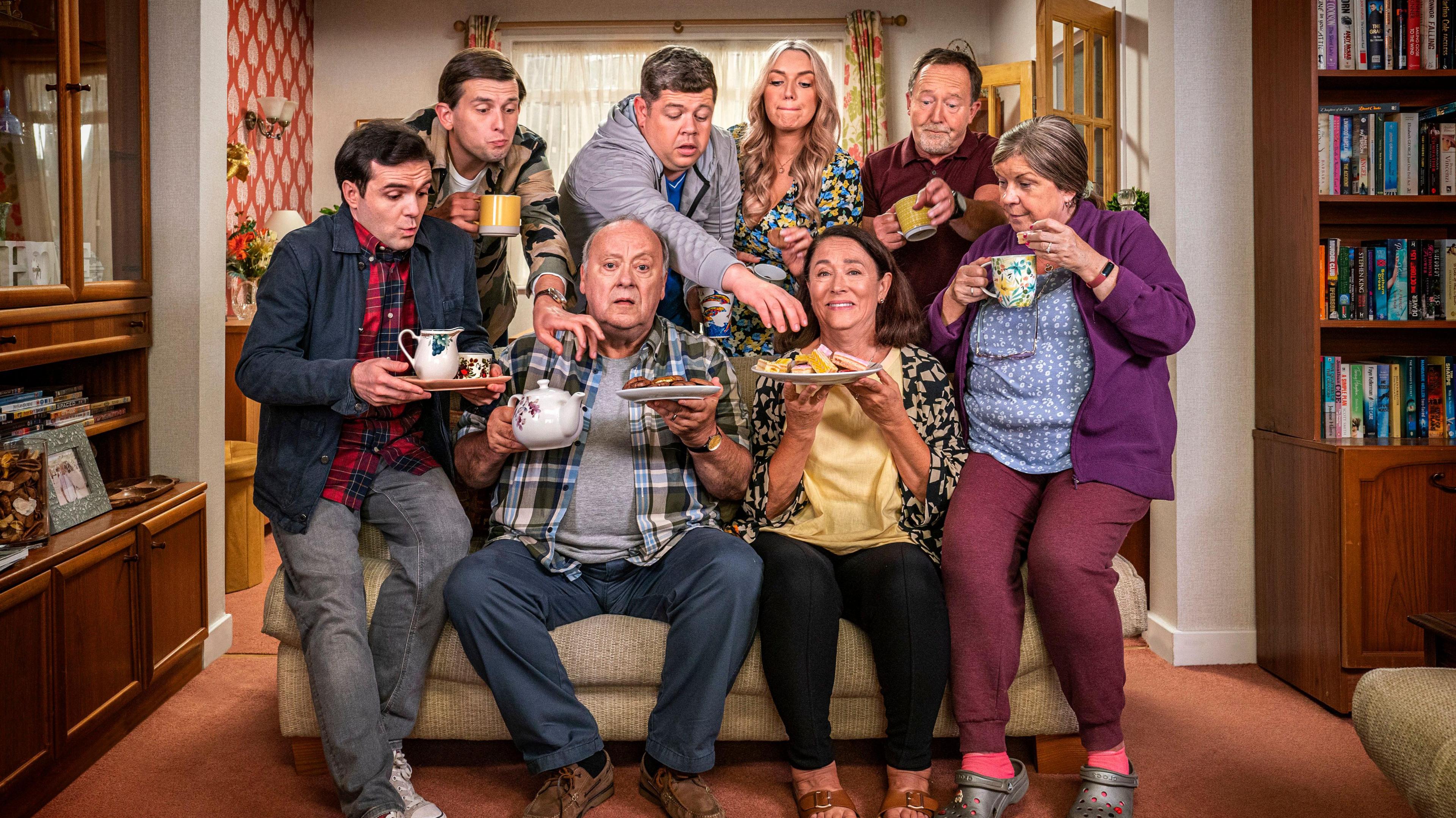 The cast of Two Doors Down crowded around a sofa in a living. Elaine C Smith - as Christine - is to the right of the group, perched on the arm of the sofa with a cup of tea and a slice of cake.