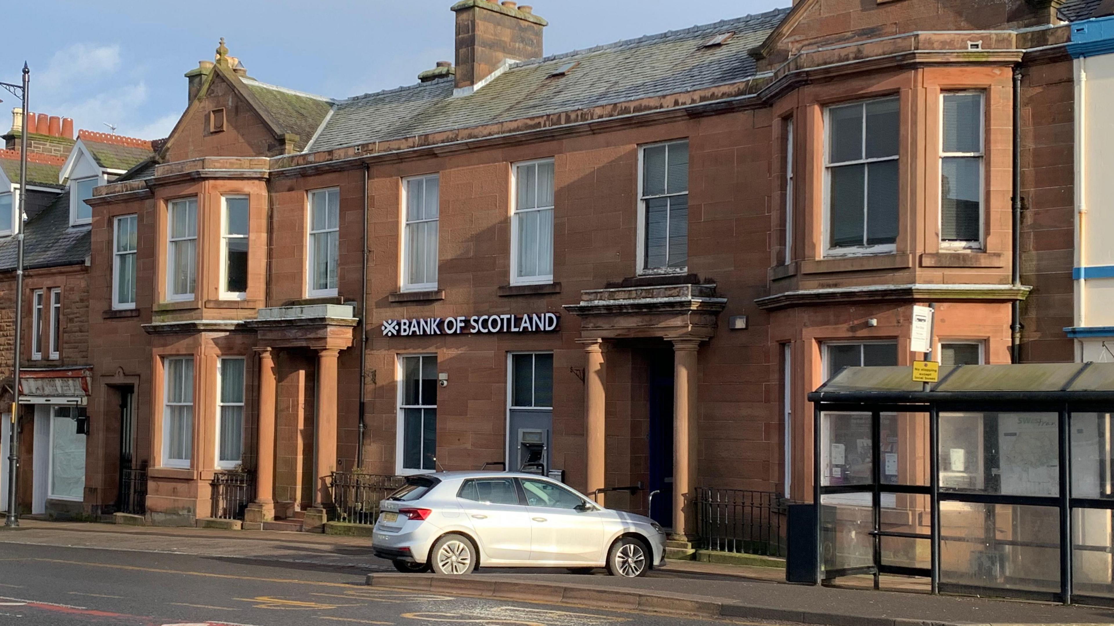 A sandstone fronted building with the Bank of Scotland name and logo on the front with a car parked outside and a bus shelter nearby