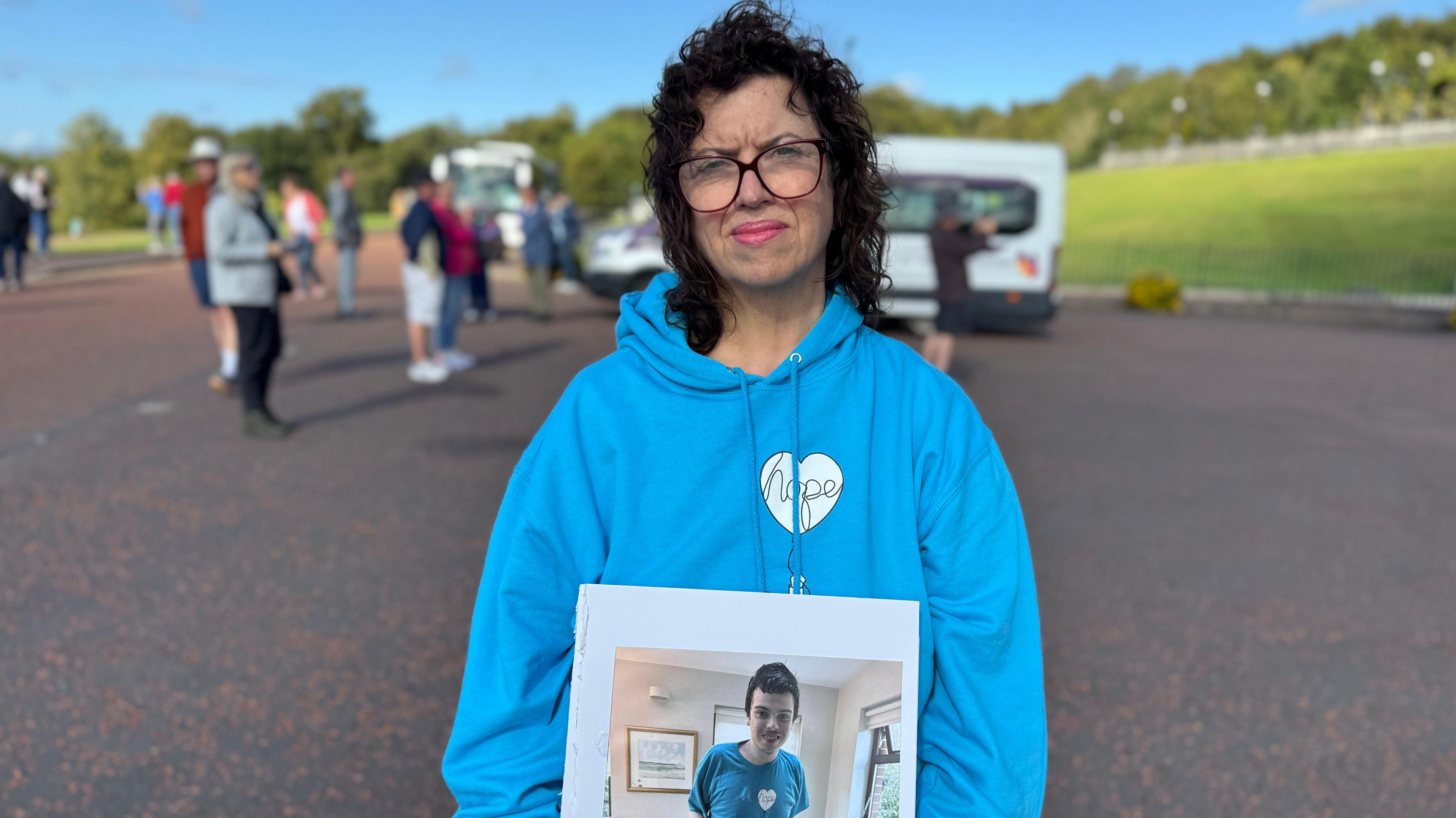 Sheena McCann is wearing a blue hoodie, and is holding a photo of a brown haired boy wearing a blue top. Behind her, out-of-focus are several people and a white van.