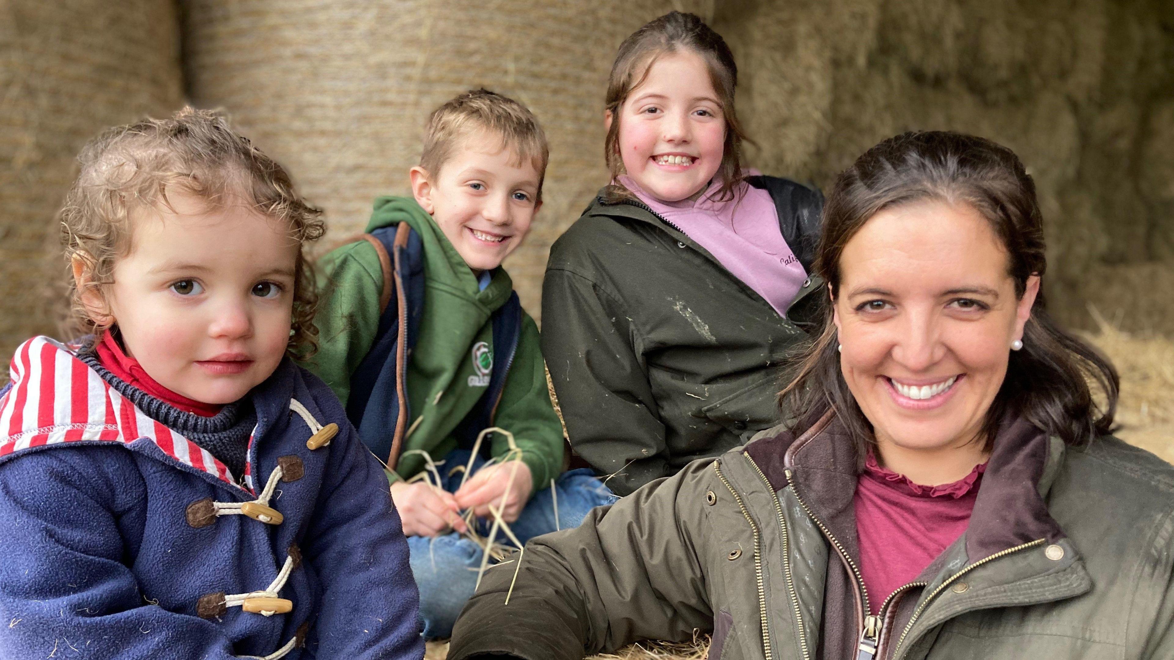 Lucy Holt looks at the camera and smiles. She has her three children with her who are sitting on hay bales and are at her shoulder level. All three are looking at the camera and the oldest two are smiling. A curly haired toddler is looking at the camera with a neutral expression.