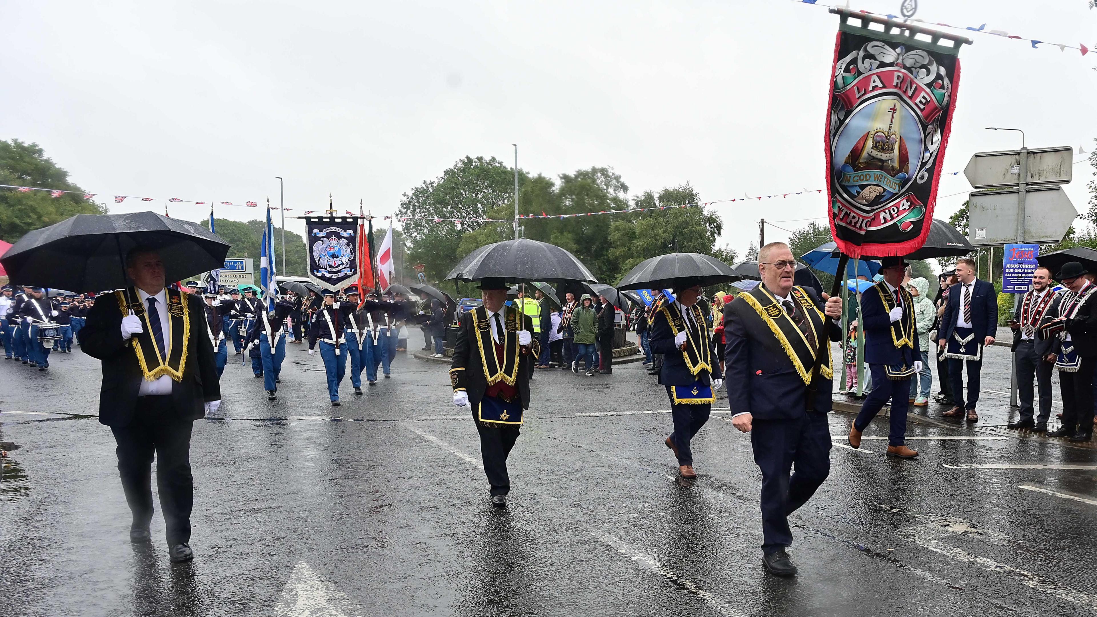 Members of the Royal Black Institution walking along a street carrying banners and flags. They are wearing dark suits and black sashes, some of them have also got black bowler hats on.