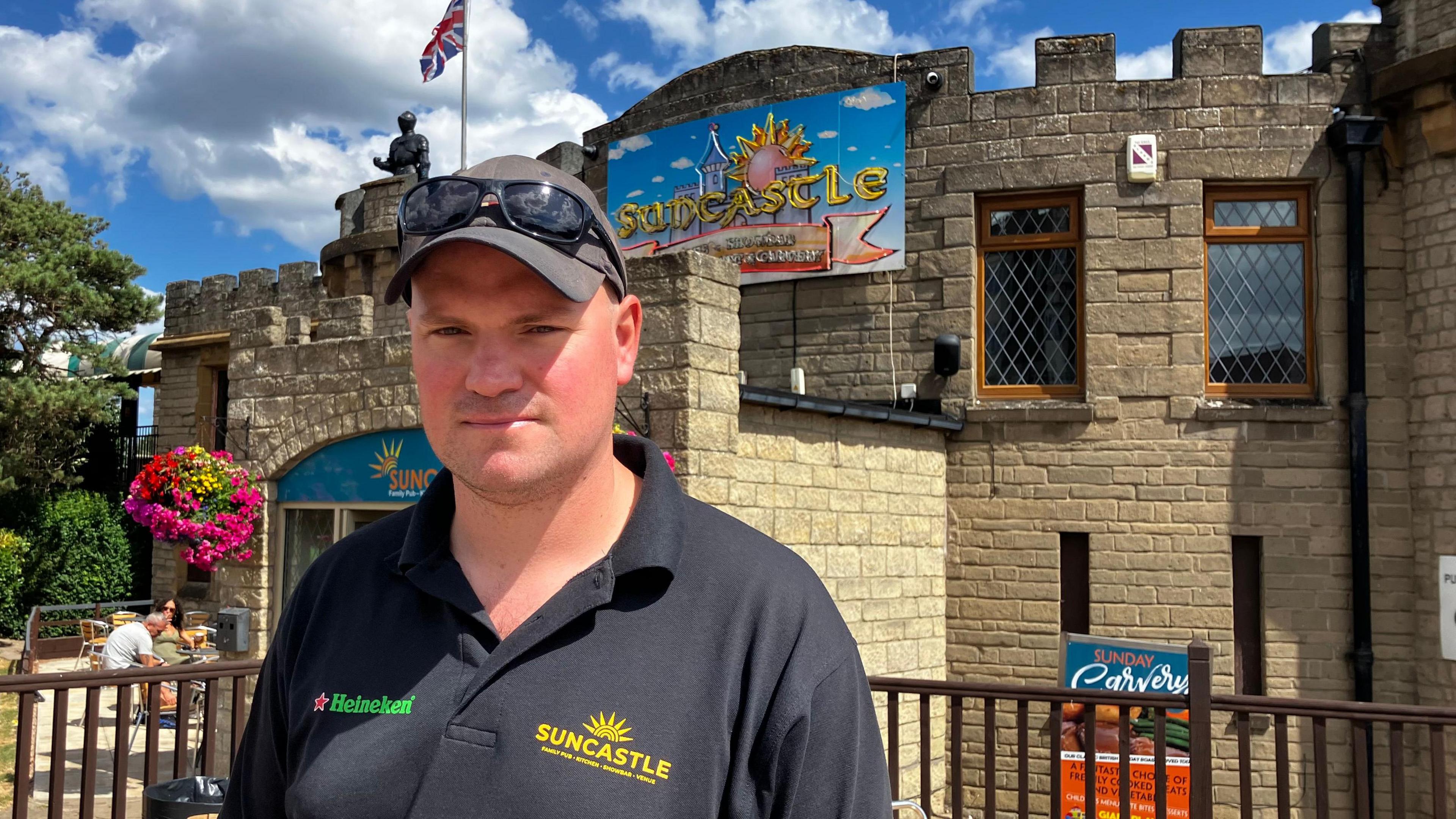 A man in a black polo shirt which says Suncastle on the left-hand side looks at the camera. He is wearing a dark baseball cap and sunglasses sit on top of the rim. He is standing in front of some chairs and tables and a building shaped like a castle is behind him.
