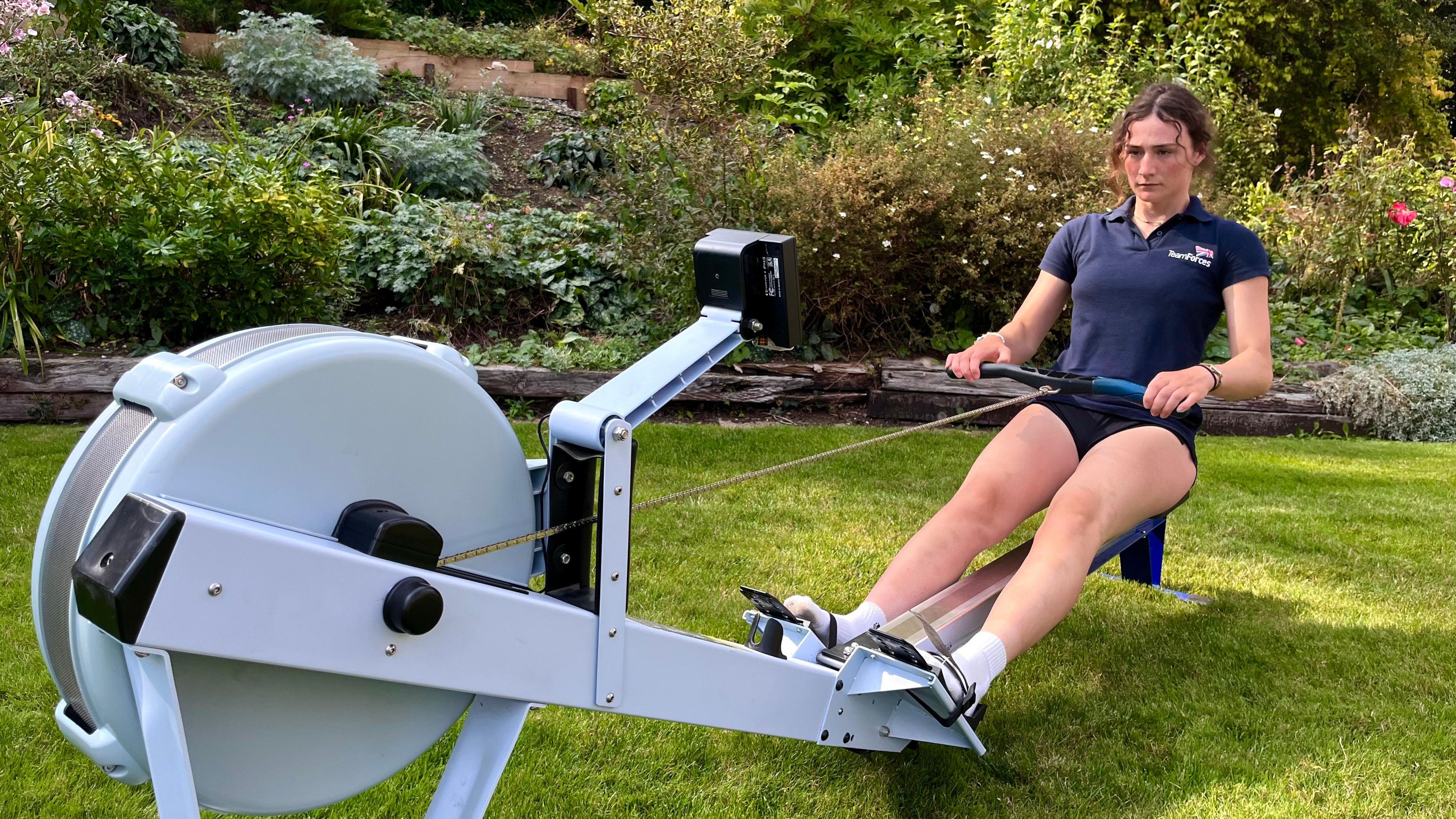 Zara working out on a rowing machine in the garden, with well-manicured flower beds behind.