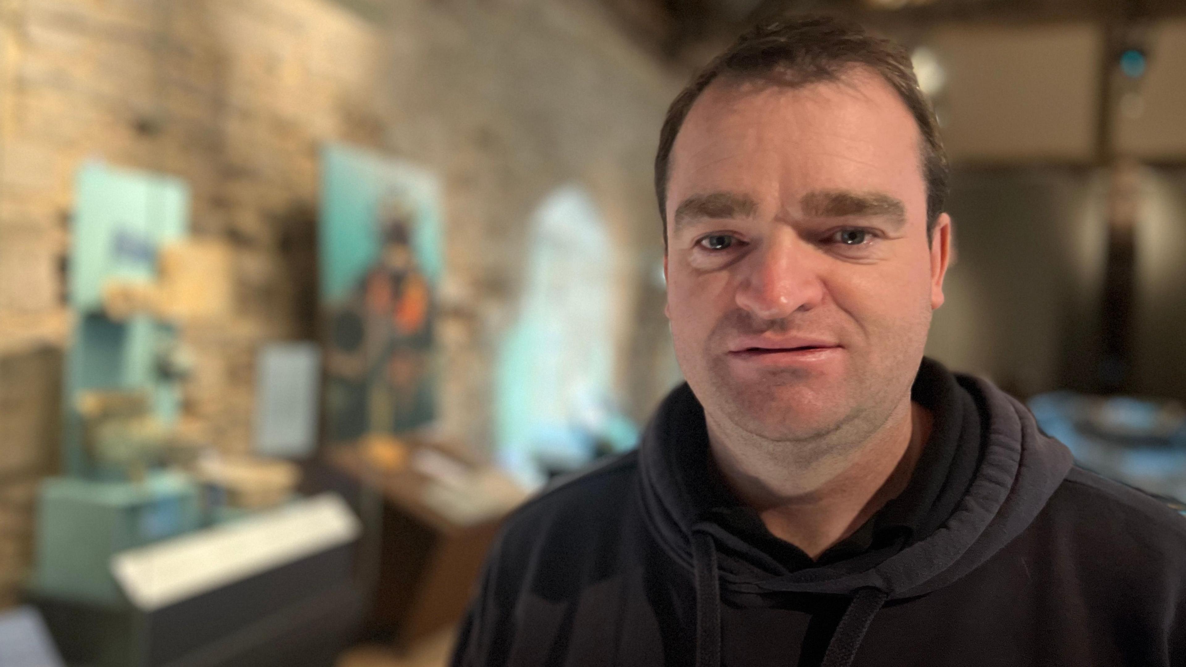Tom Cousins is wearing a black hoodie and is looking at the camera. He is standing in Poole Museum where out of focus in the background the finds from the dive can be seen on display. 