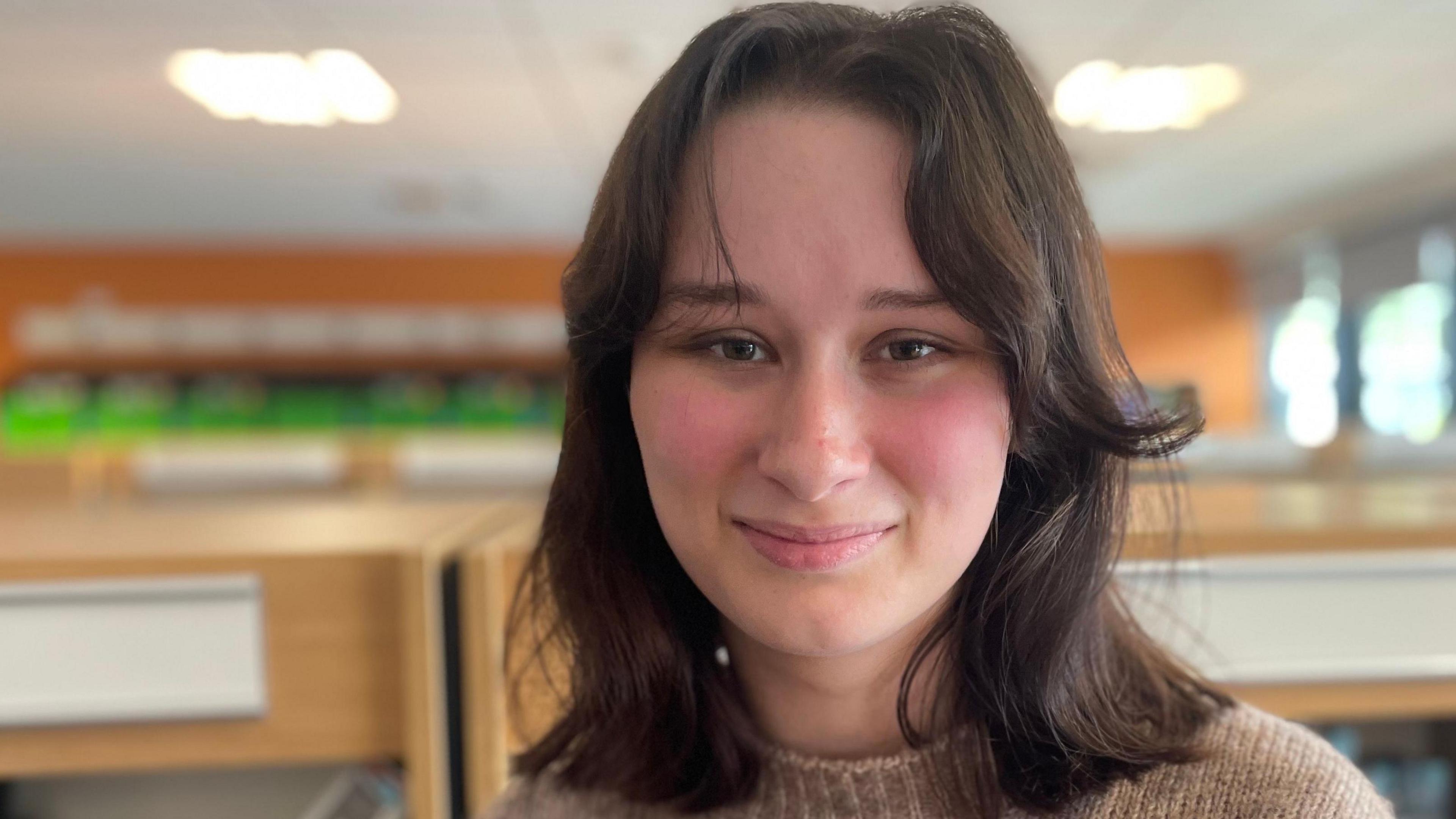 Maddie, 18, who has mid-length dark hair, and is wearing a light coloured jumper, smiling at the camera in front of bookshelves