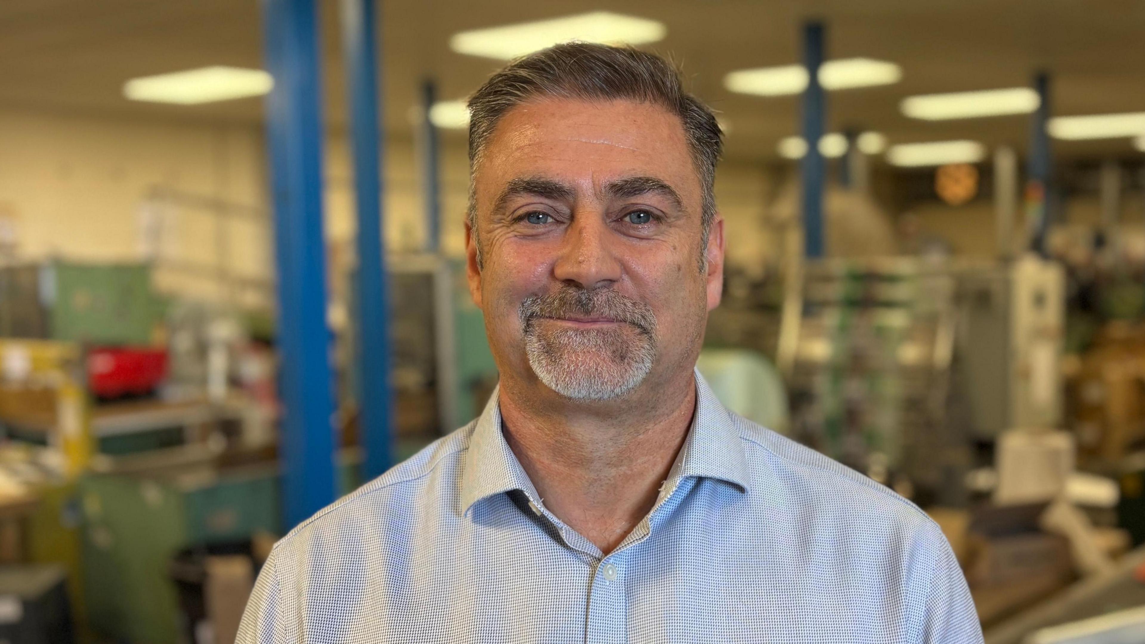 Stuart Searle looking direct to camera, standing inside an industrial-looking area. He is smirking slightly and is wearing a blue, tightly checked shirt, with its collar open. He has a greying goatee bear, blue eyes, and short dark hair, which is parted on one side. The background of the image is out of focus, but there is machinery behind Stuart, blue pillars that appear to run from floor to ceiling, and fluorescent lighting.  