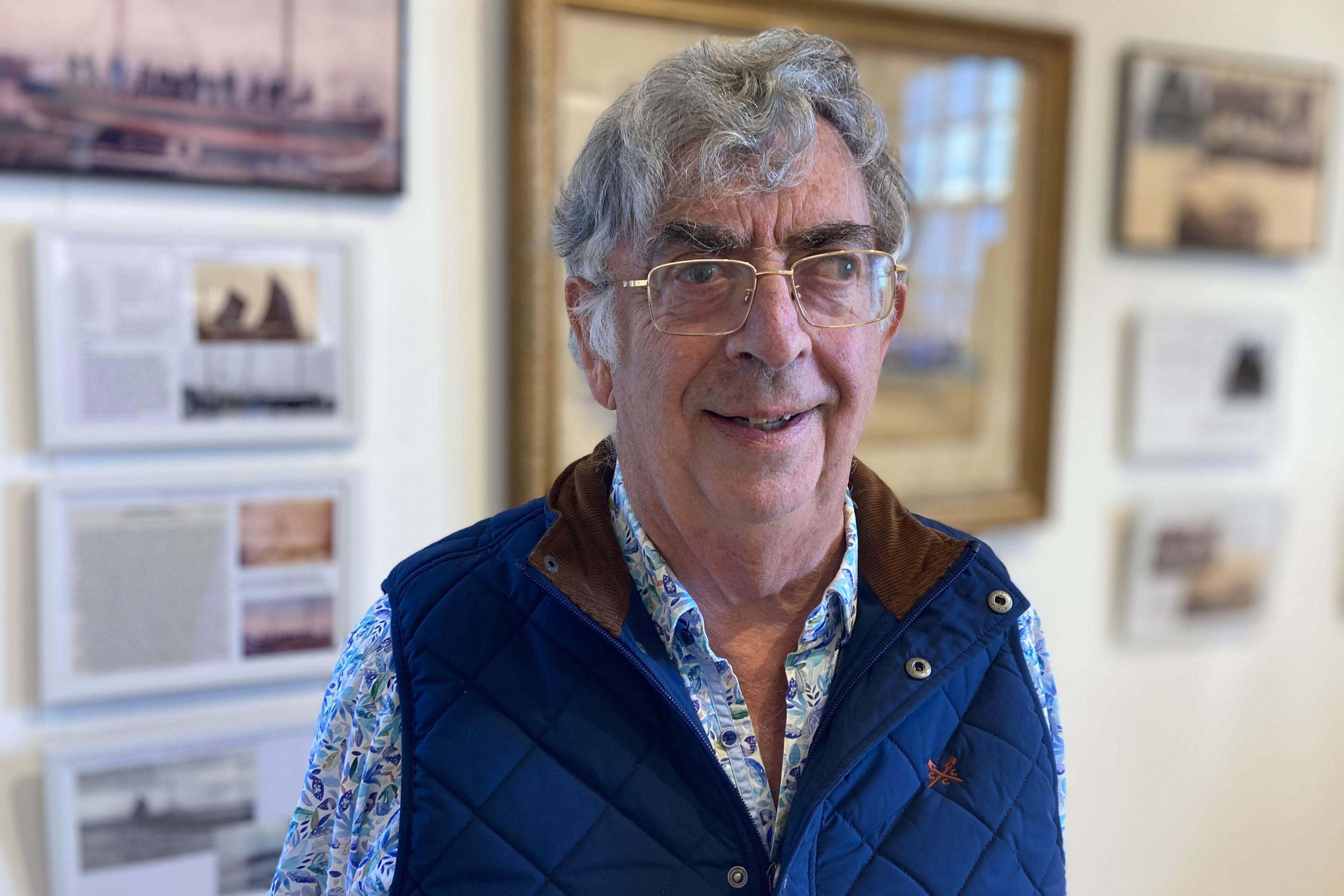 Tony Bone wearing a blue jacket stood in front of a wall, the wall has pictures on.