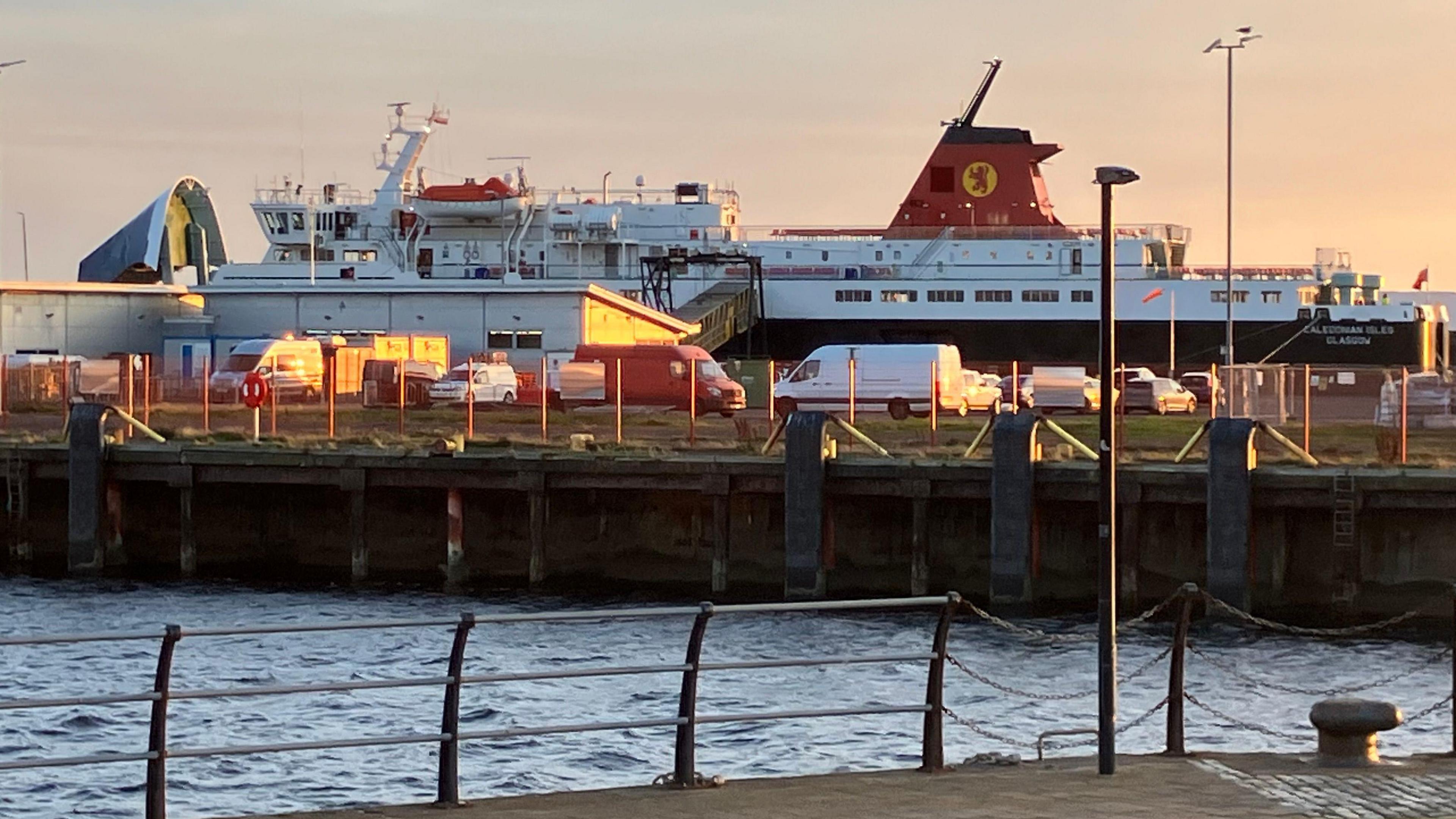A large passenger ferry docked in a harbour. Parked vehicles can be seen in the foreground