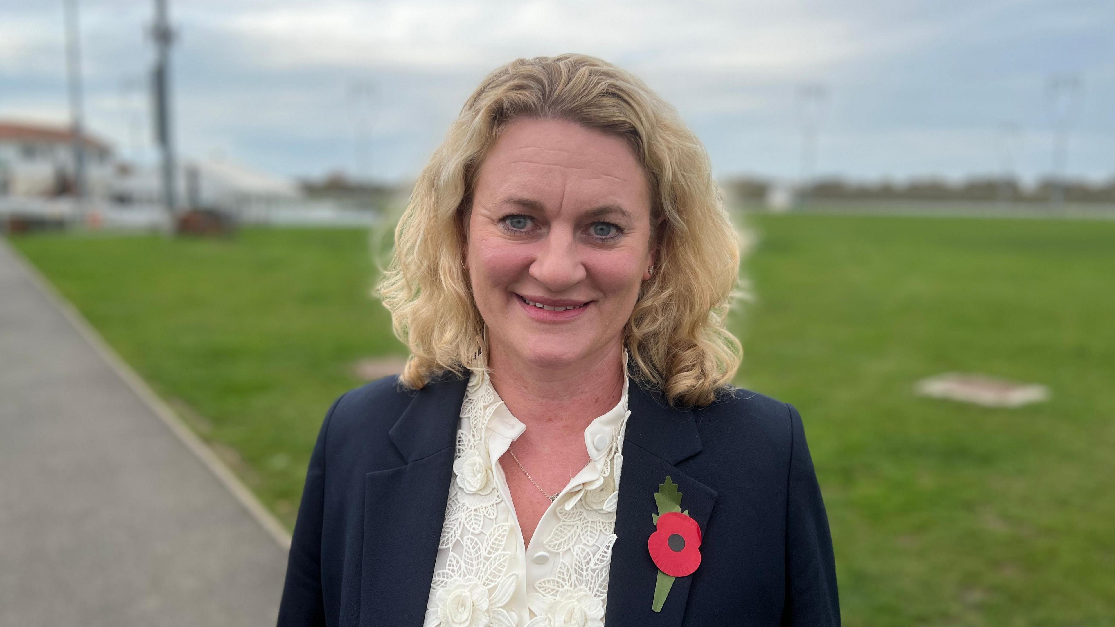 Louise McKinlay stands on a pathway in front of a section of green grass. She has shoulder length blonde hair that is slightly curled. She wears a navy suit jacket with a poppy on one side of her chest and a cream blouse underneath.