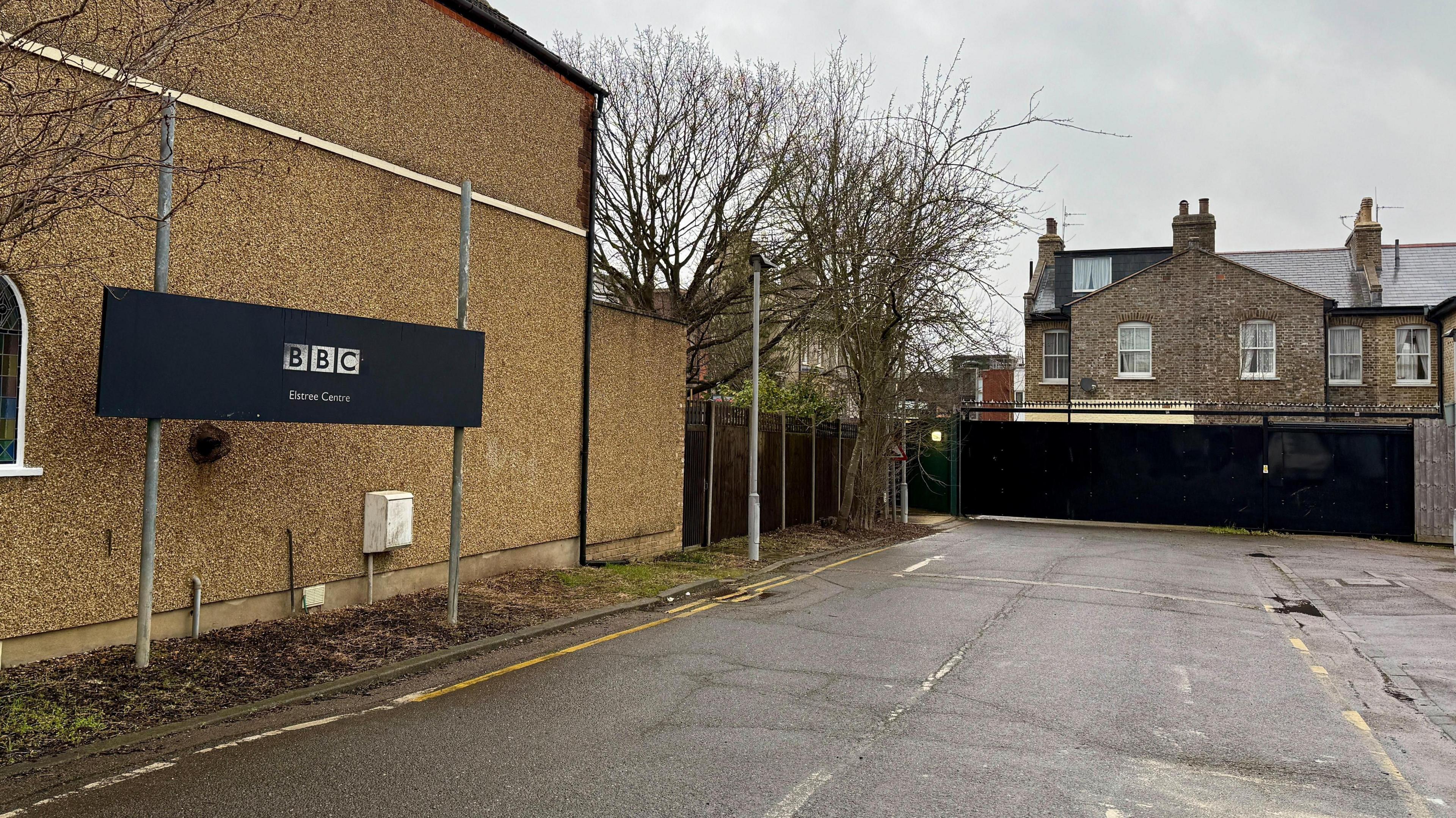 The access road that runs towards the EastEnders set. A large black sign reads "BBC Elstree Centre". Houses can be seen on the other side of a black security gate